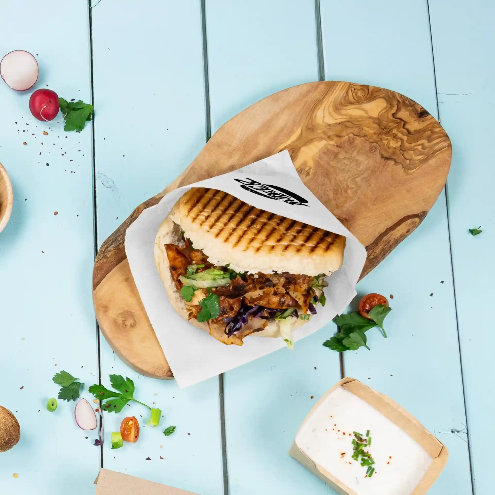 Image of a sandwich on a wooden board with a printed sleeve, surrounded by fresh vegetables and a small container of sauce.