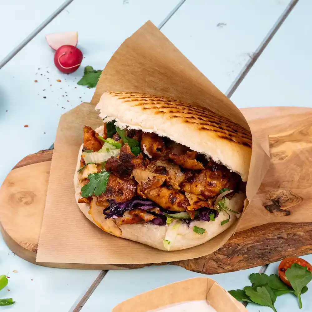 A filled pita sandwich with grilled chicken, lettuce, purple cabbage, and cilantro on a wooden board.