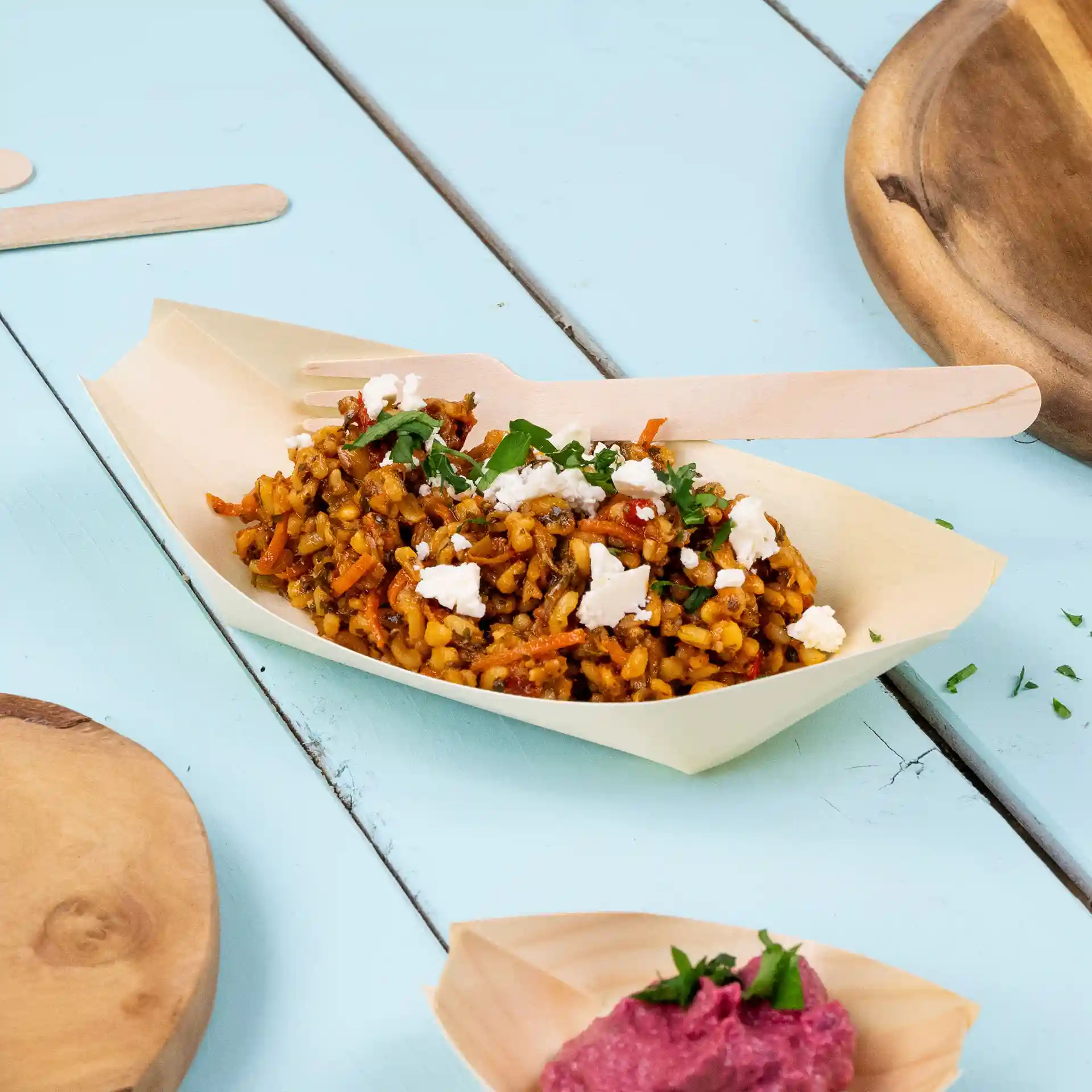 Wooden boat-shaped dish with rice, vegetables, and feta cheese, accompanied by a wooden fork on a blue surface.