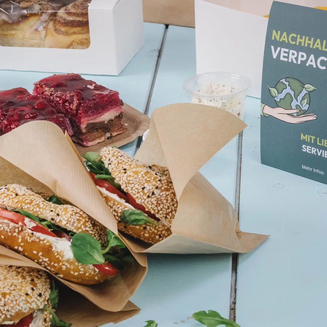 Three sesame seed buns filled with lettuce and tomato, a dessert with jelly, and a sustainability-themed sign on a blue table.