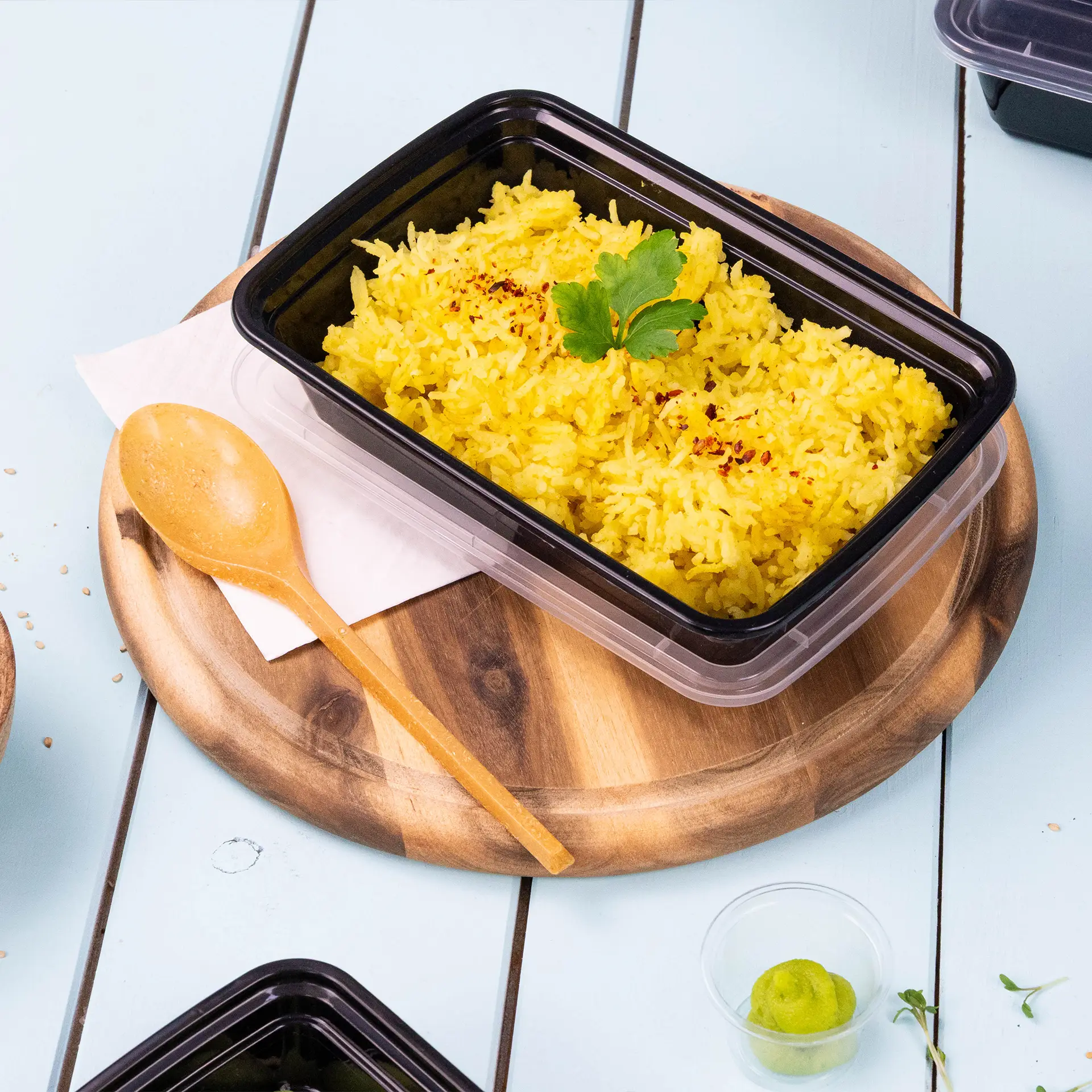 One black container with yellow rice topped with a green garnish, placed on a wooden board with a reusable brown spoon nearby.