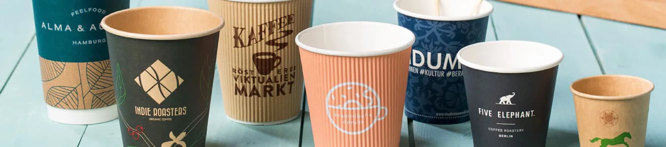 A collection of various paper coffee cups featuring unique designs and logos, arranged on a light wooden surface.