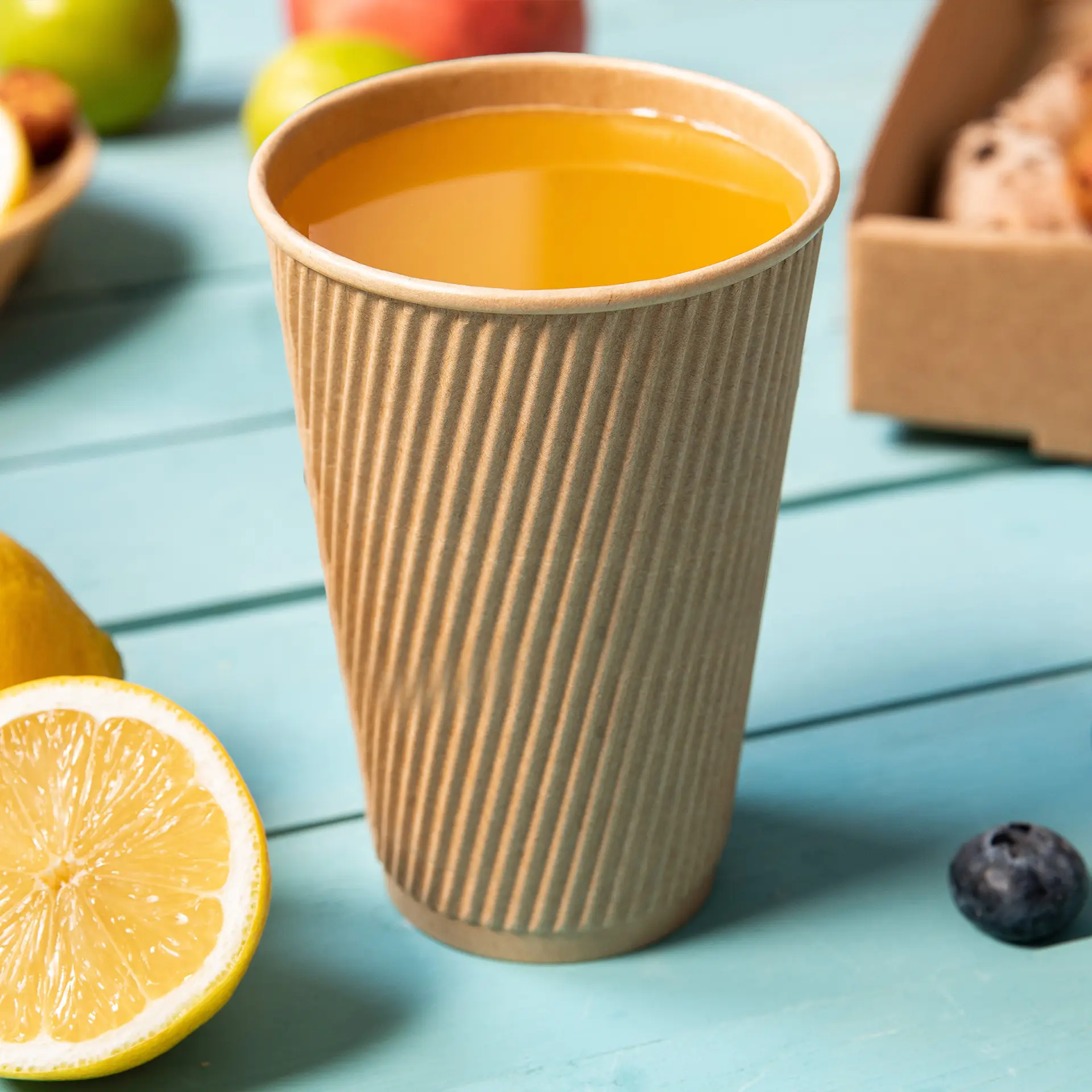 Brown triple-walled disposable coffee cup filled with orange drink, surrounded by fruit on a turquoise background.
