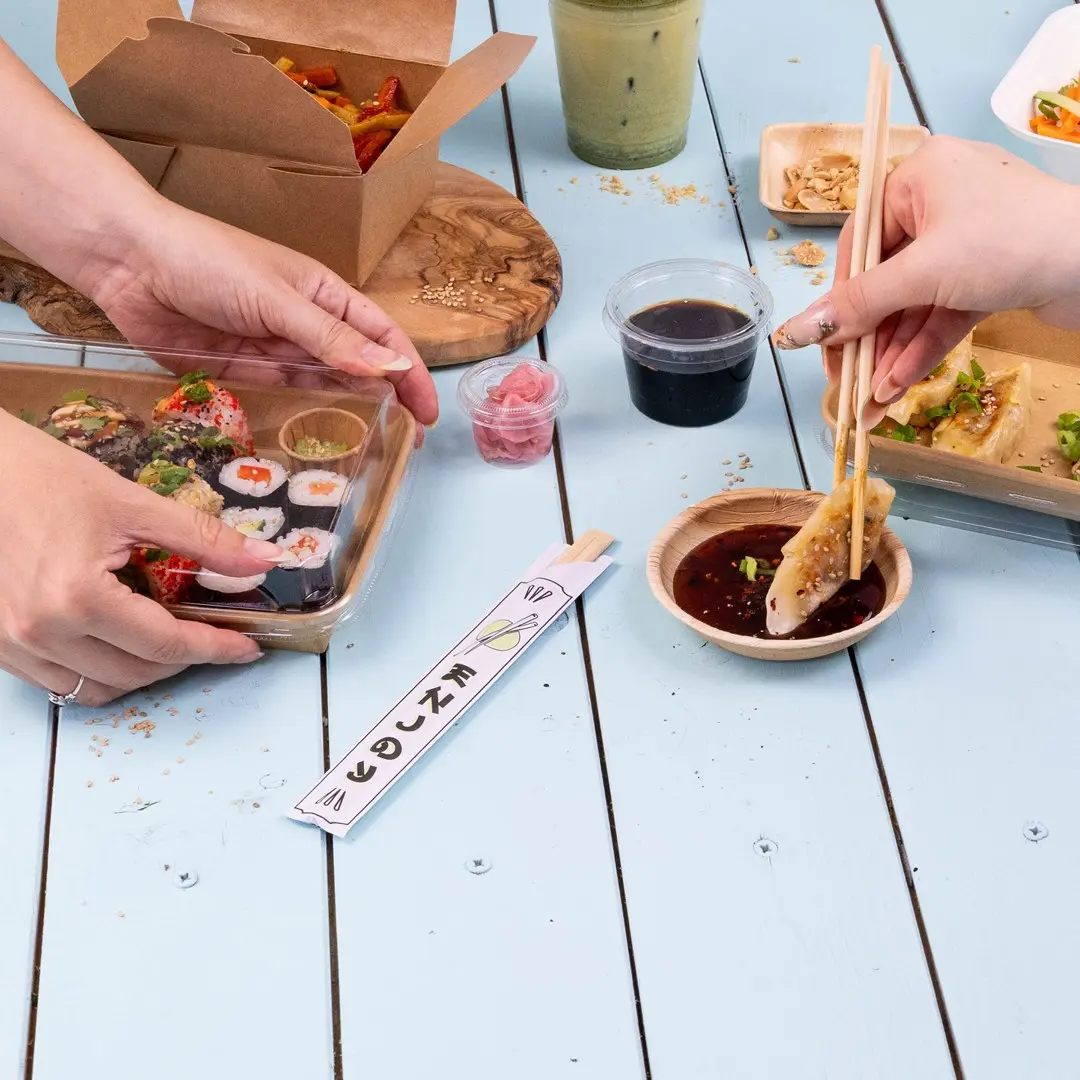 Hands holding sushi in a clear container, with dipping sauce, pickled ginger, and chopsticks on a blue table.