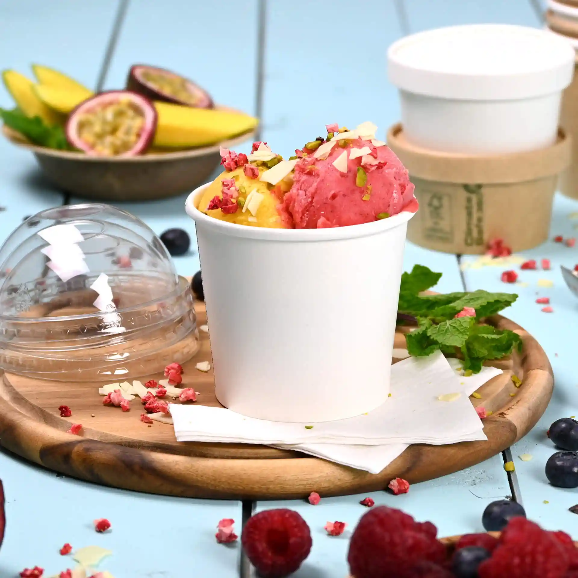 White paper ice cream tub with pink and yellow sorbet topped with nuts and dried fruit, placed on a wooden tray.