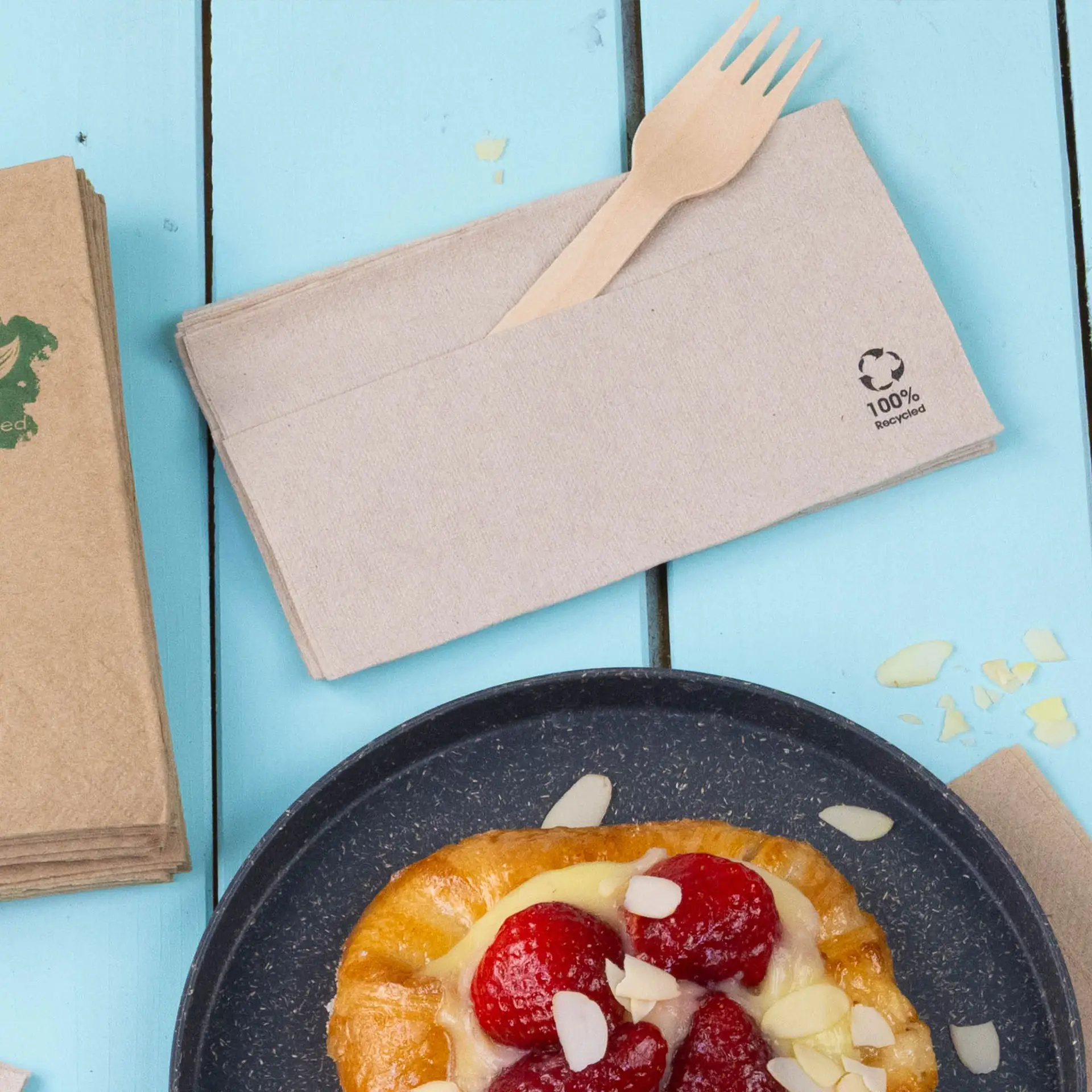 Recycled paper napkin with a wooden fork, placed beside a pastry on a blue surface.