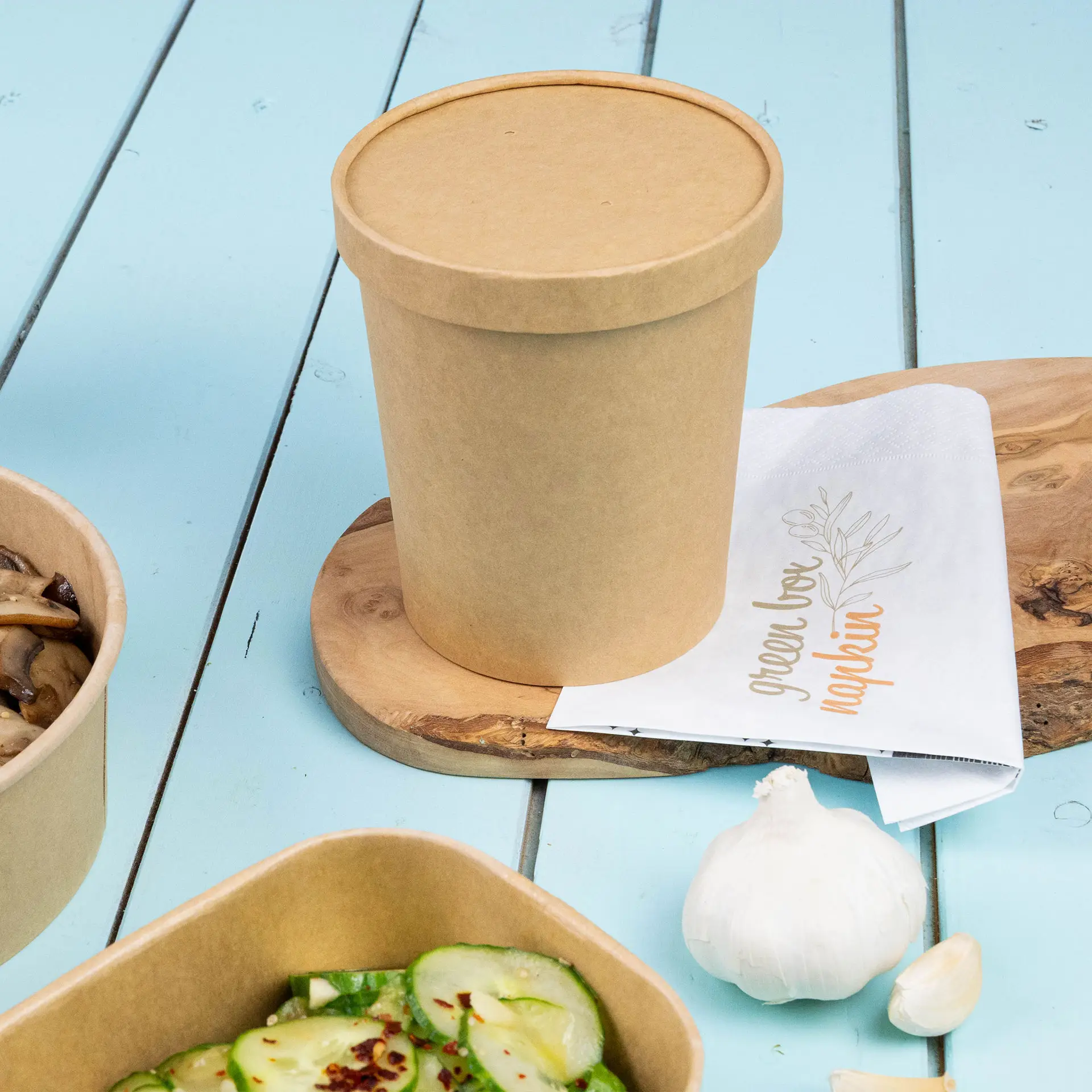 Brown cardboard soup cup with a lid, placed on a wooden board, alongside a napkin and food containers.