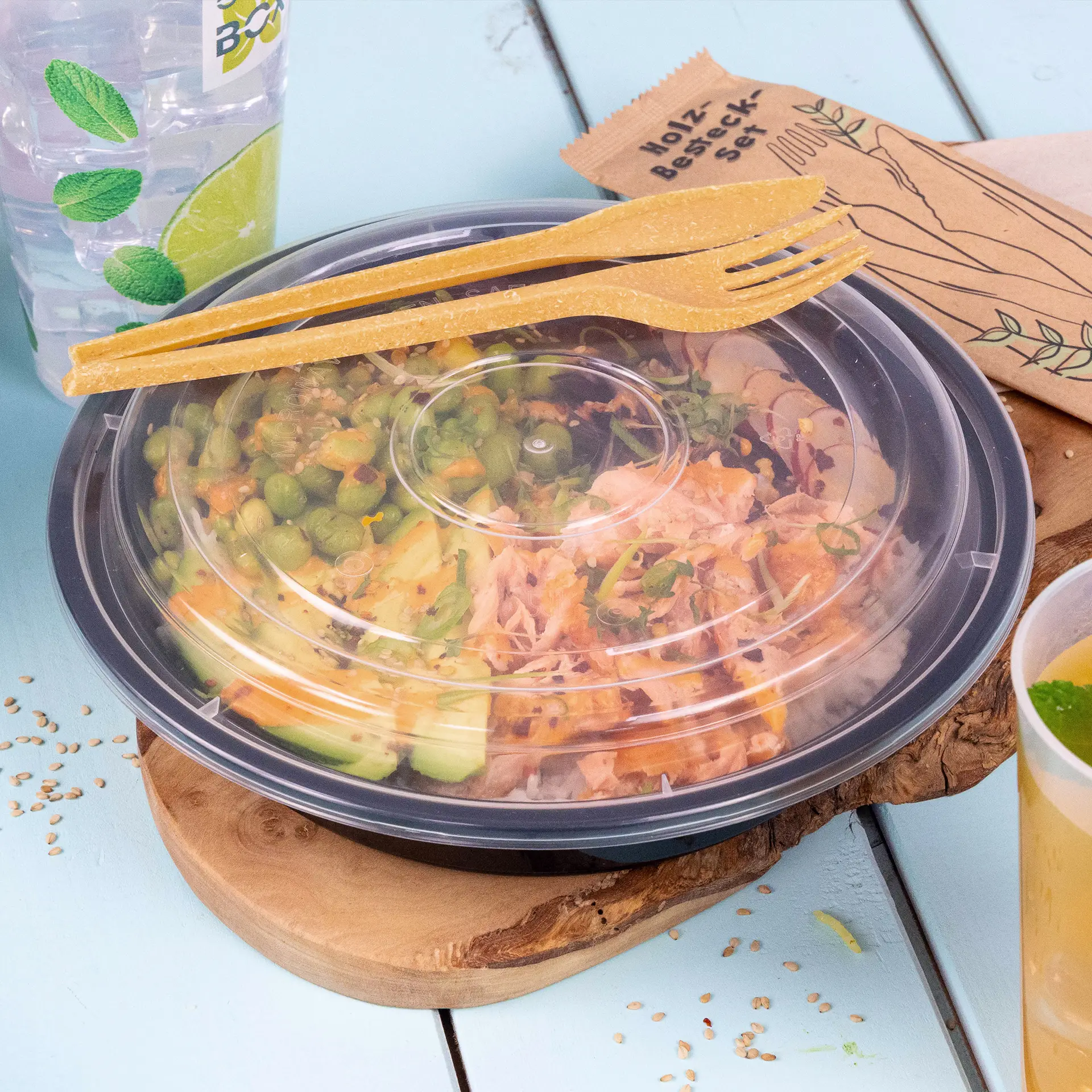 One clear plastic food container filled with colorful salad ingredients, topped with a caramel reusable knife, and a beverage in the background.