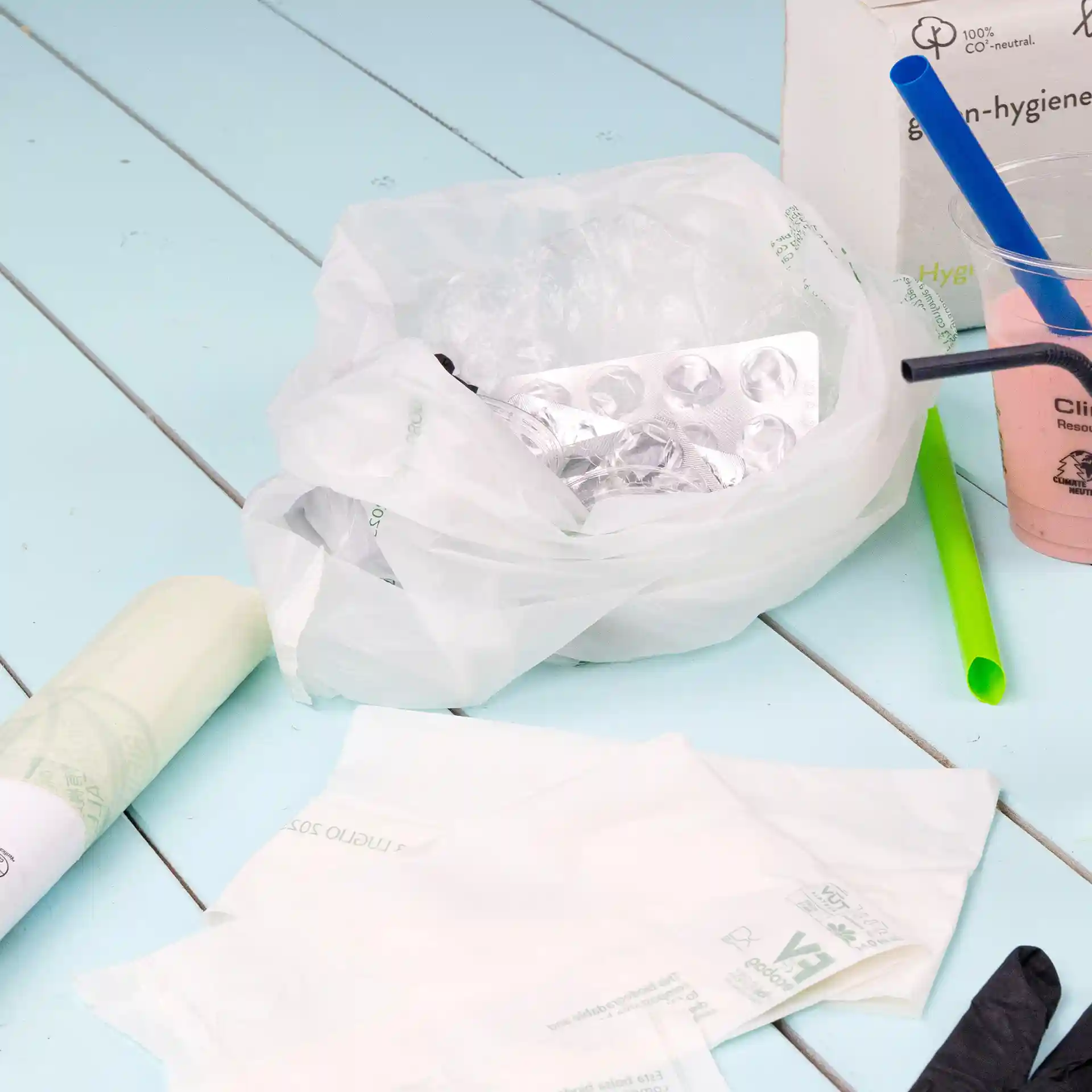 Transparent bio bin bag with contents including a blister pack, plastic straws, and disposable items on a blue surface.