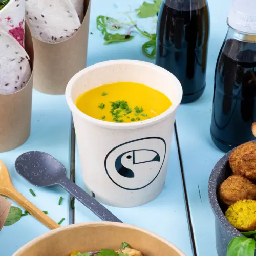 Soup cup with yellow soup and green garnishes, surrounded by food containers and drinks on a blue surface.