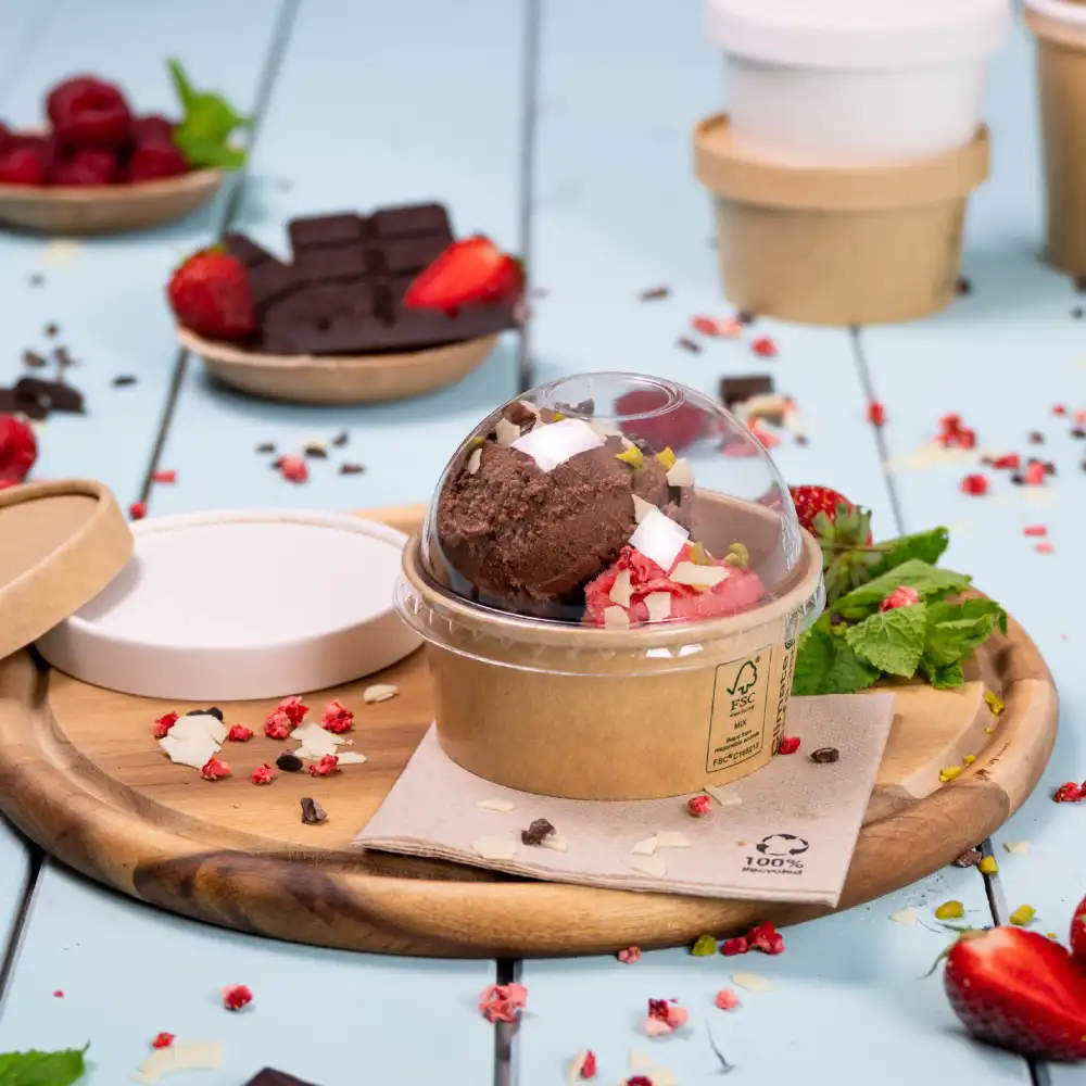 Ice cream in a transparent cup with a dome lid, placed on a wooden tray with chocolate, strawberries, and raspberries in the background.