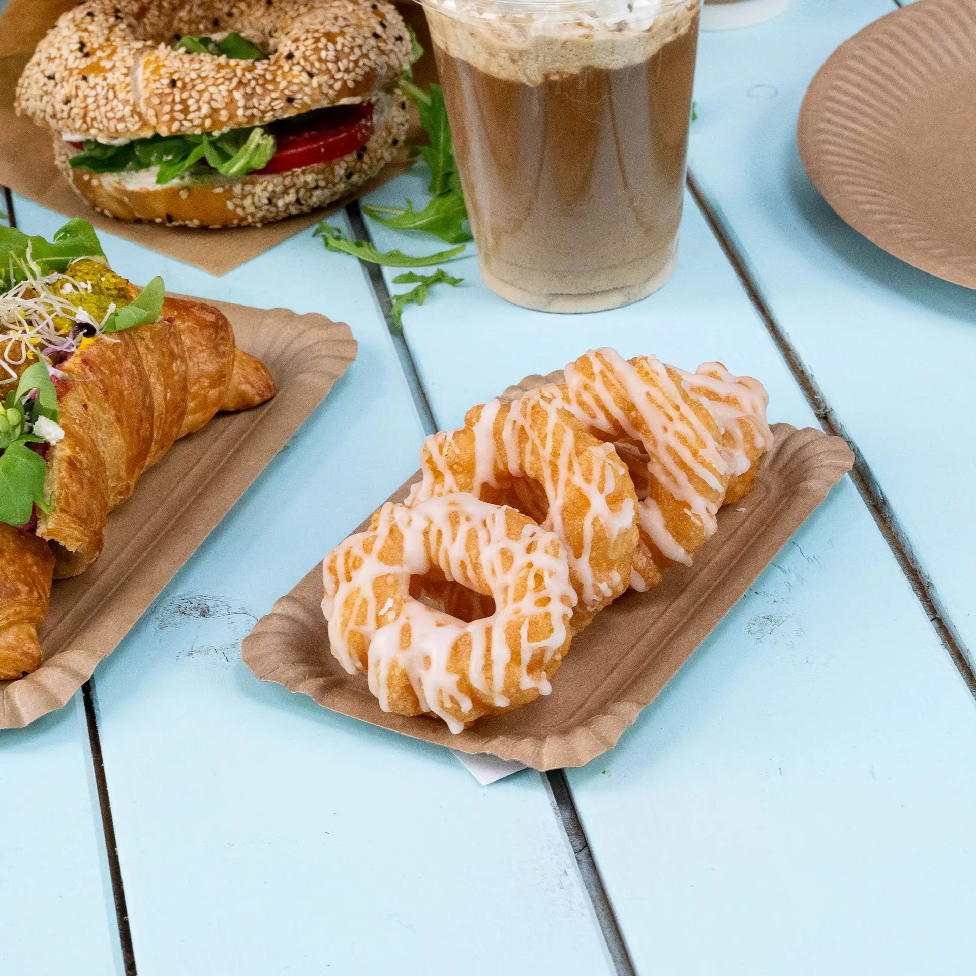 Three frosted doughnuts on a rectangular brown cardboard plate; a croissant and a bagel are also visible in the background.