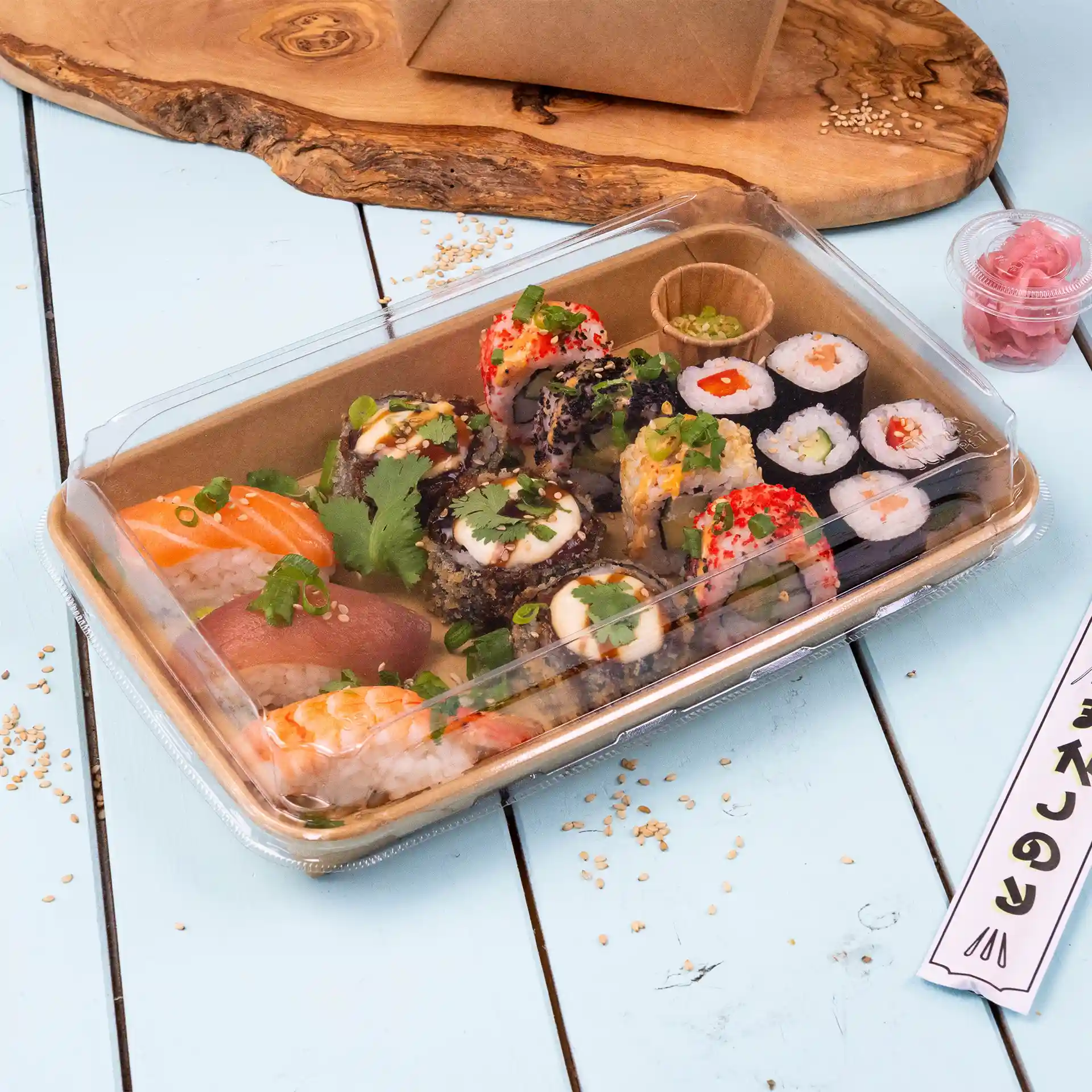 Transparent sushi box lid with assorted sushi rolls, including salmon, tuna, and garnishes, on a light blue surface.