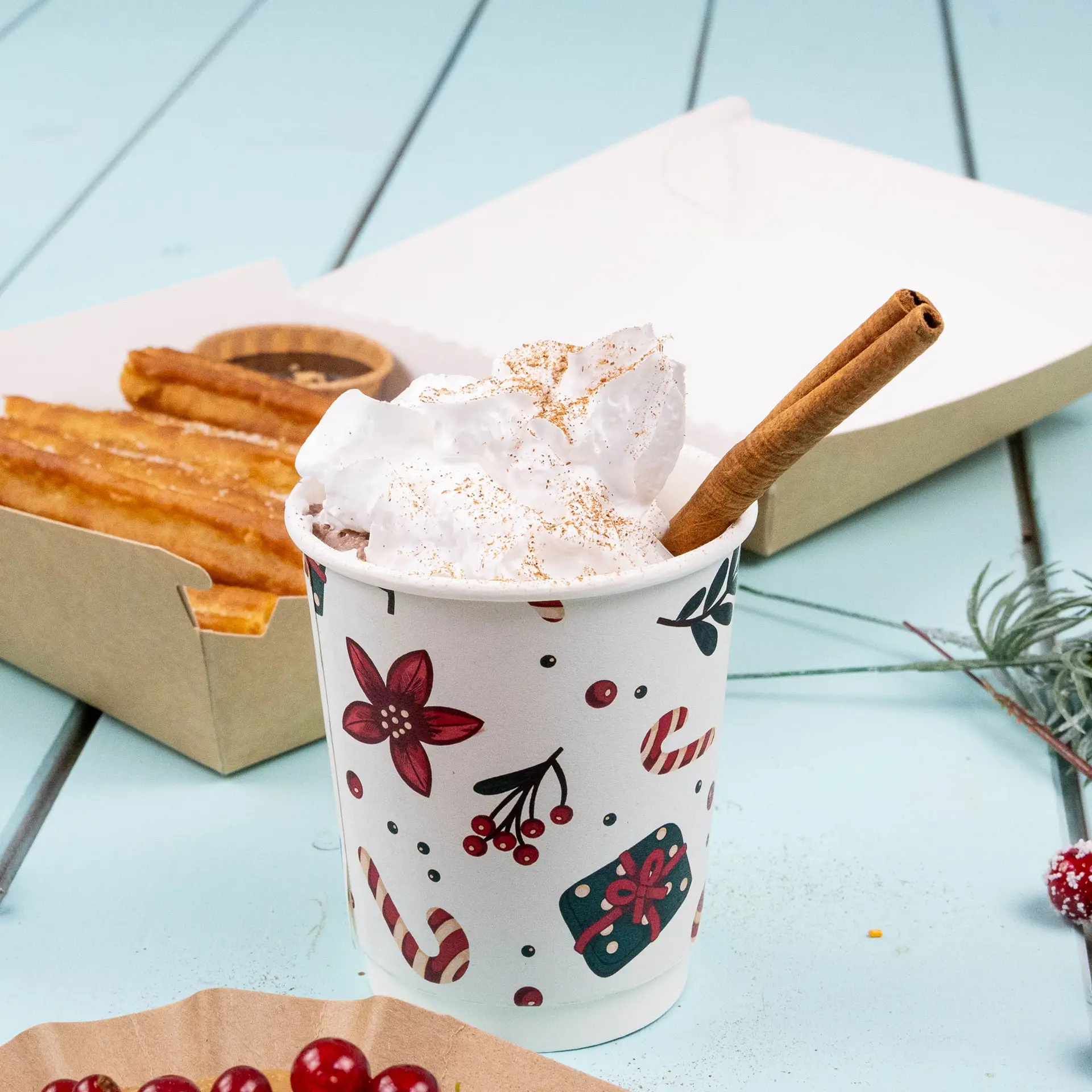 Double-wall coffee cup with winter design, filled with whipped cream and a cinnamon stick, alongside churros in a box.