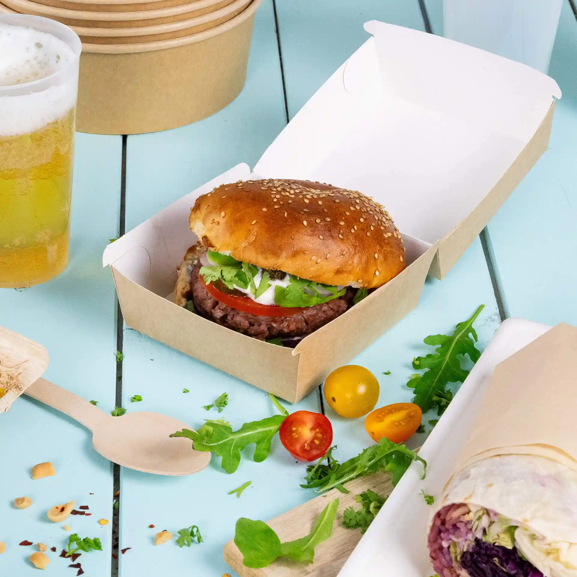 Brown cardboard takeaway burger box containing a sesame seed bun with lettuce, tomato, and sauce, on a blue surface.