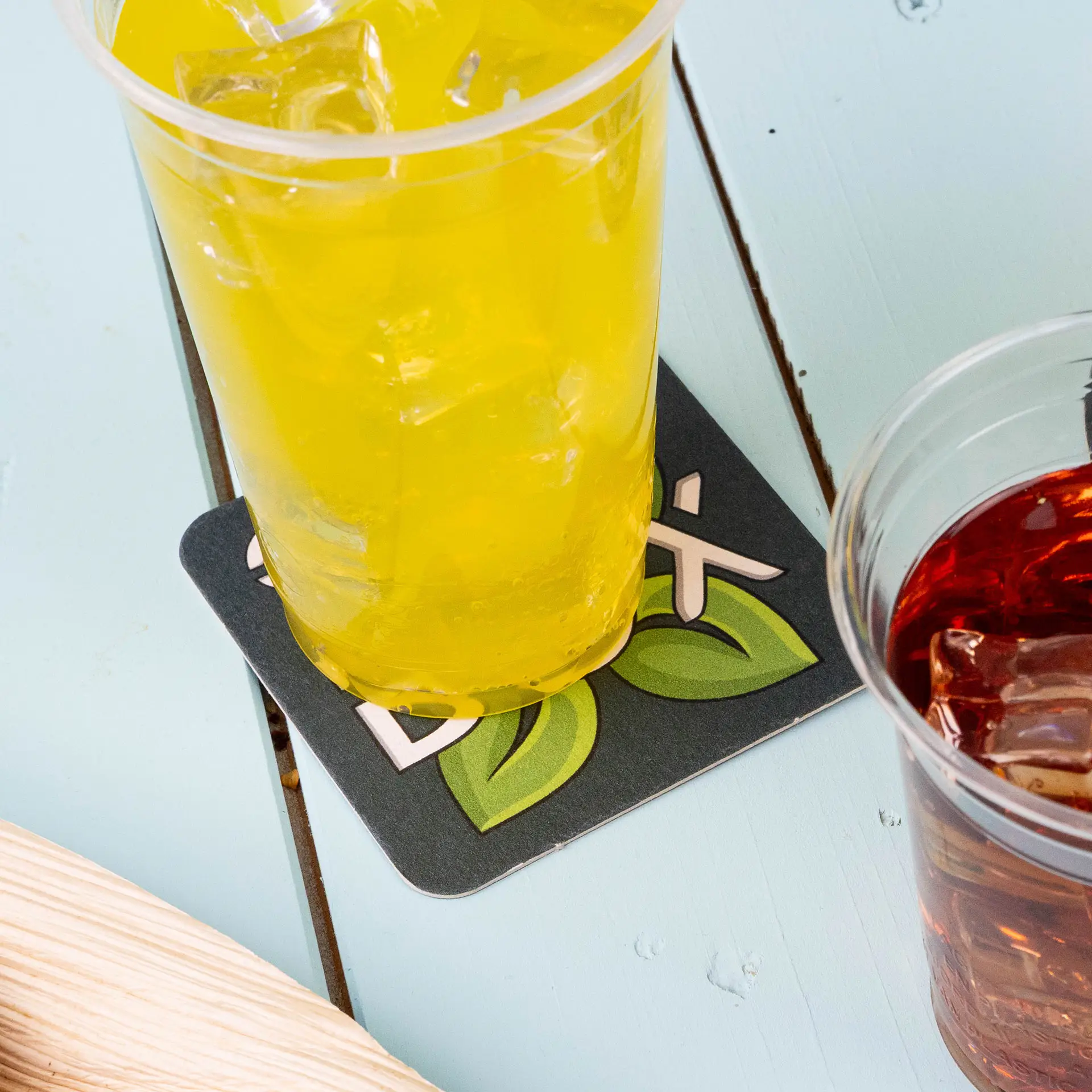 A yellow drink in a plastic cup sits on a printed square beer coaster with leaf designs and the word 'Box'.