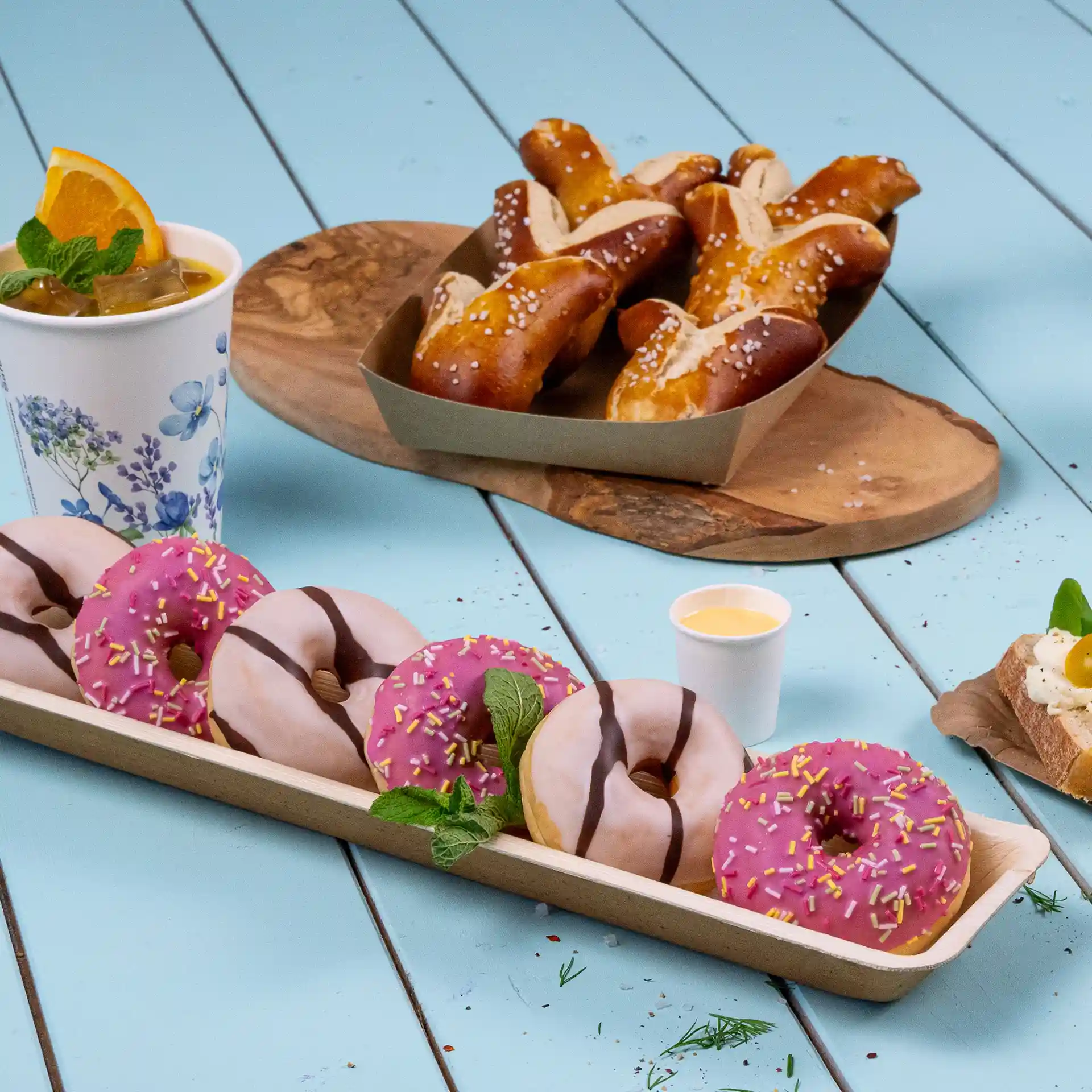Rectangular palm leaf tray with several donuts, a drink, and pretzels on a blue surface.