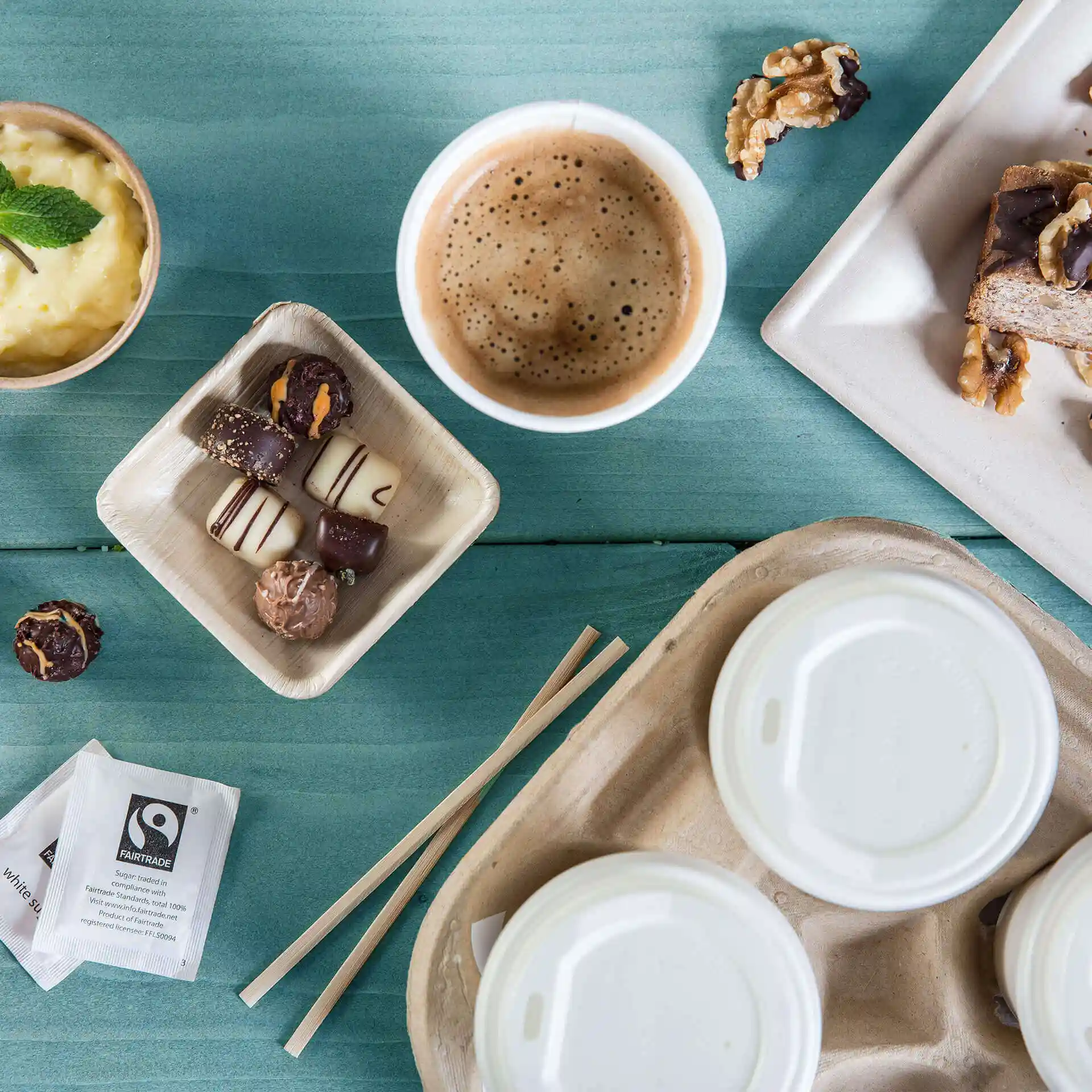 Image of a coffee cup, assorted chocolates in a tray, dessert, and a takeout container on a blue wooden surface.
