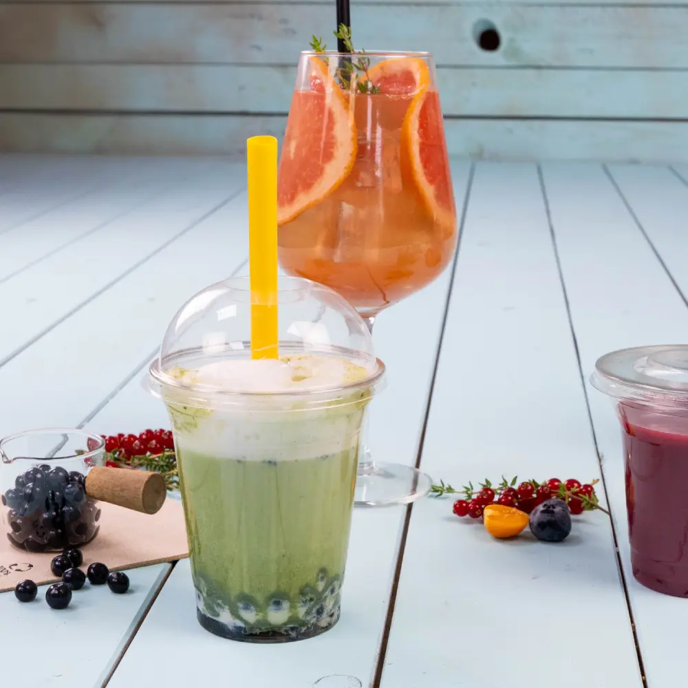 A green beverage with a yellow straw in a clear cup, with blue tapioca pearls at the bottom; a colorful cocktail with grapefruit slices in a glass behind it.