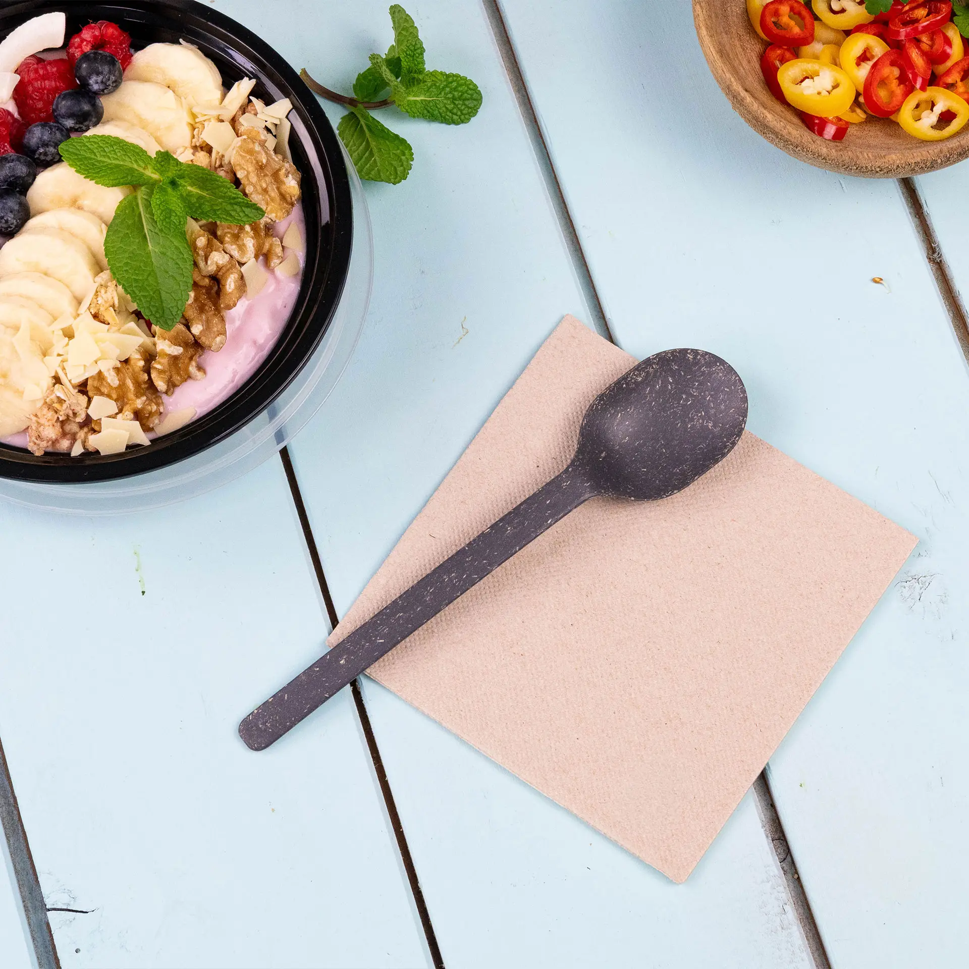 Image of a black spoon on a beige napkin, with a bowl of yogurt topped with fruits and nuts in the background.