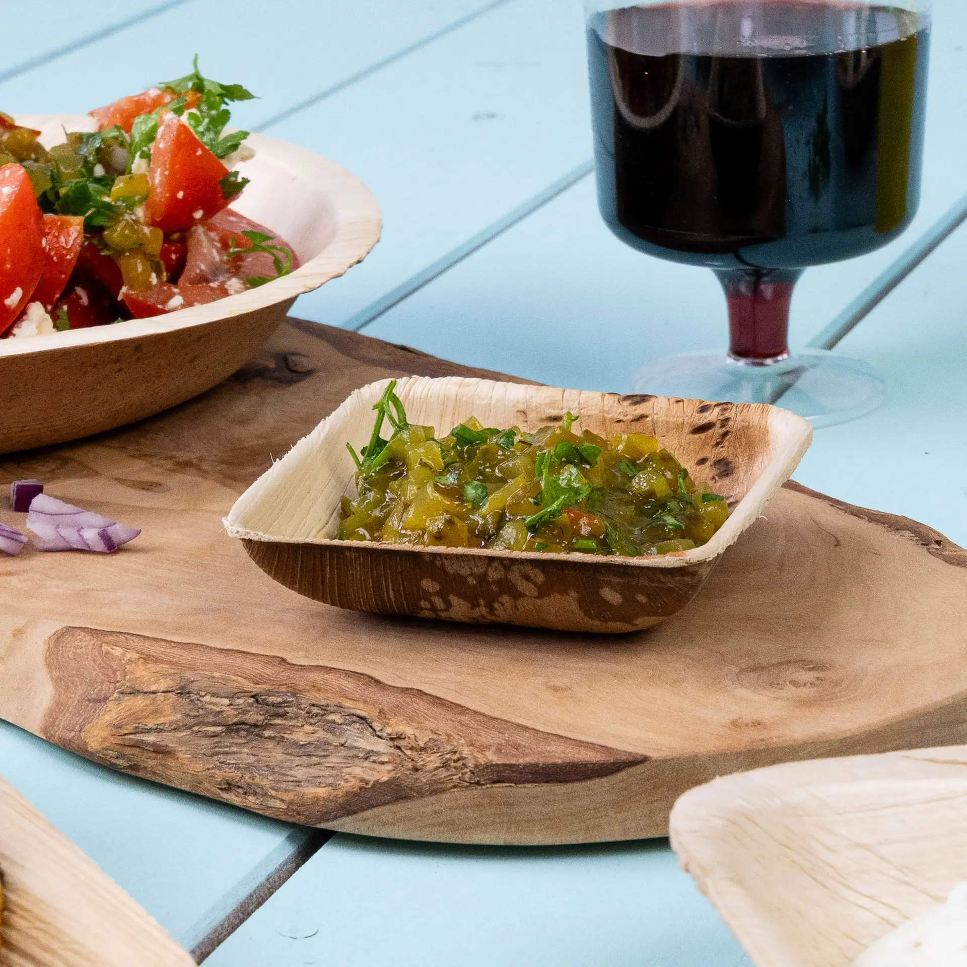 Mini square palm leaf bowl with marinated green olives; several similar bowls are visible in the background.
