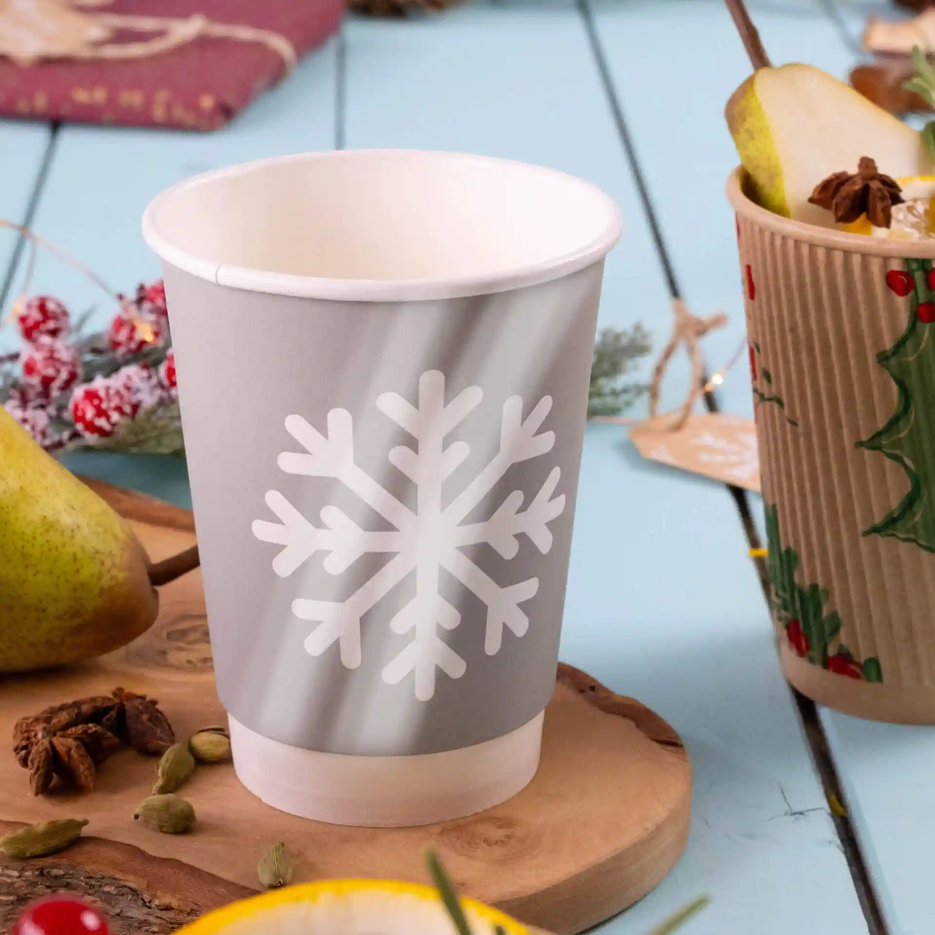 Gray paper cup with a white snowflake design, double-walled, placed on a wooden surface with a pear and spices nearby.