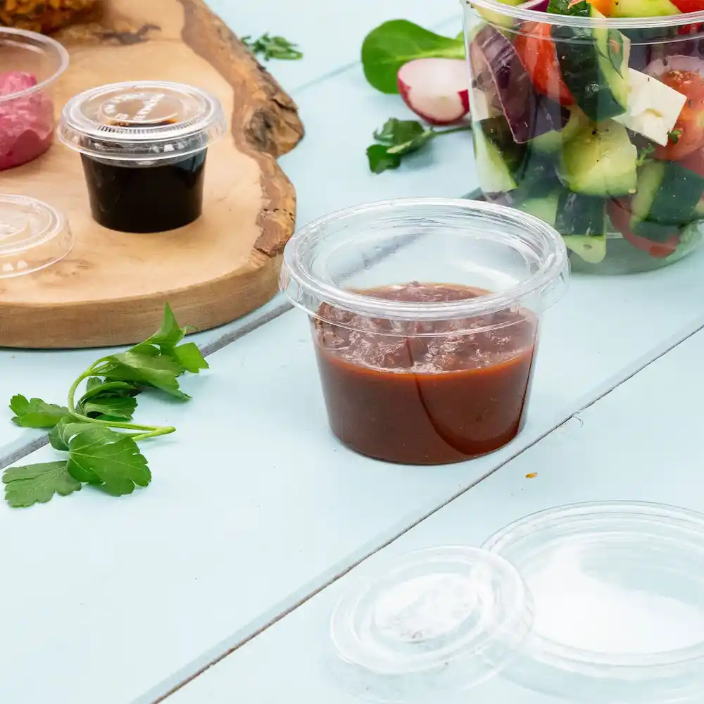 Clear portion container with reddish sauce, alongside green herbs and other food items on a light blue surface.