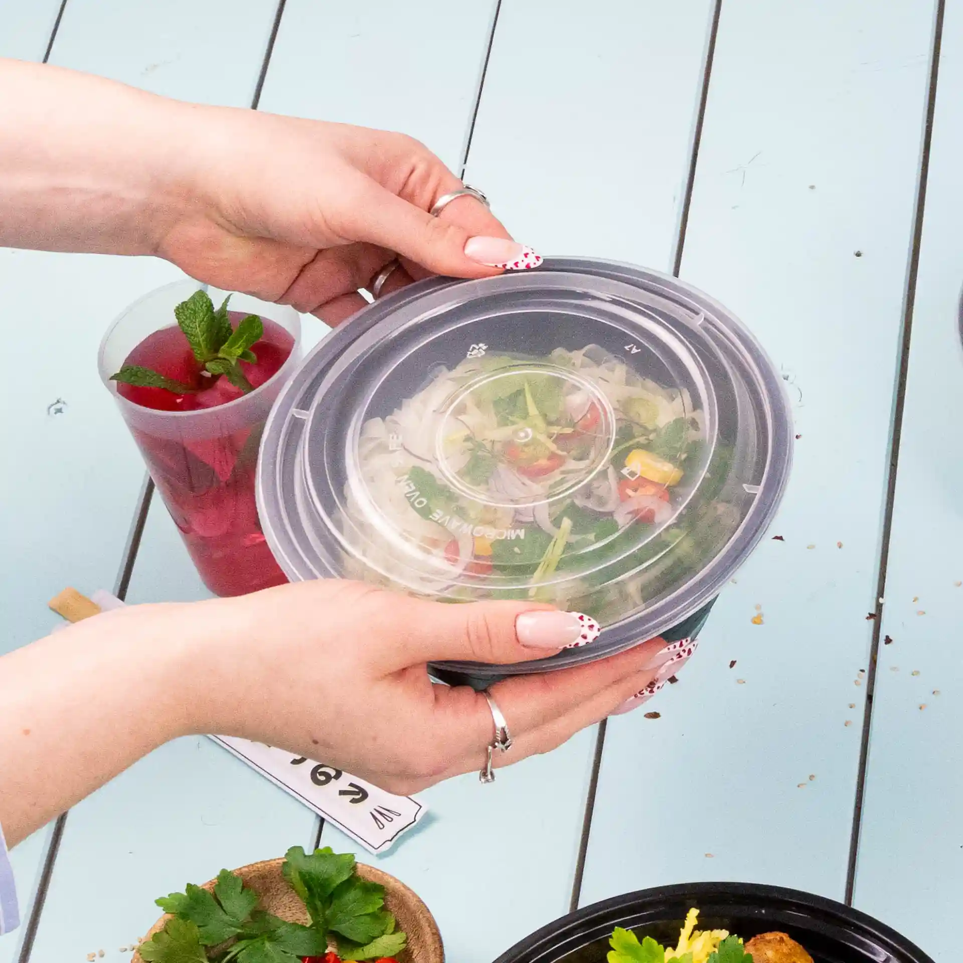 A person holding a round, transparent reusable lid made of PP above a bowl of salad on a blue table.