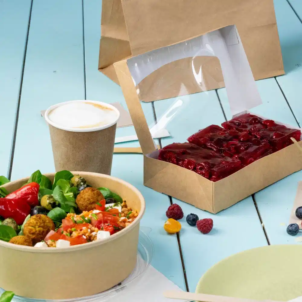 A takeaway meal setup featuring a salad in a bowl, a coffee cup, and a dessert in a transparent box on a wooden table.