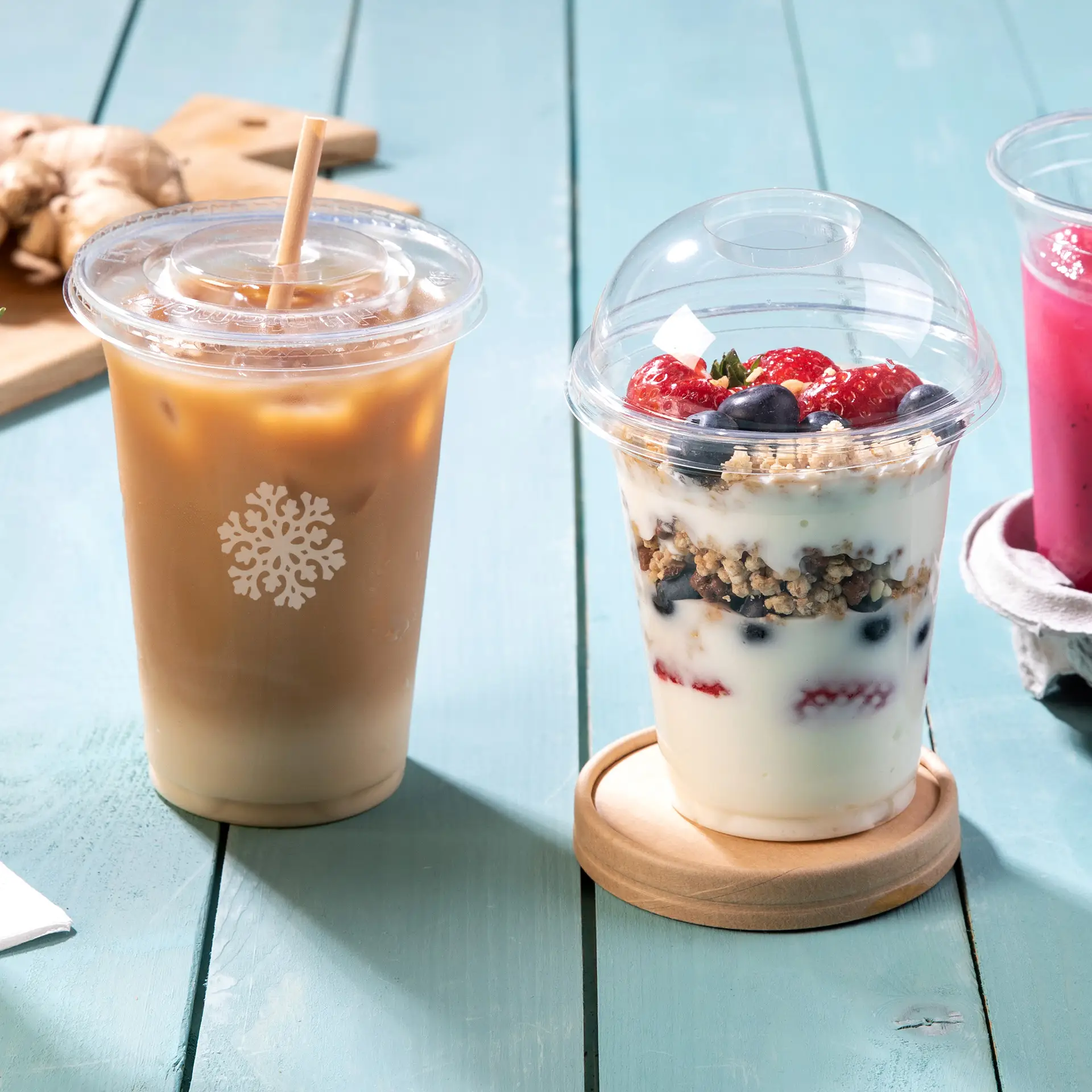 Three clear PLA cups for cold drinks: an iced coffee cup, a parfait with berries, and a pink beverage, set on a wooden table.