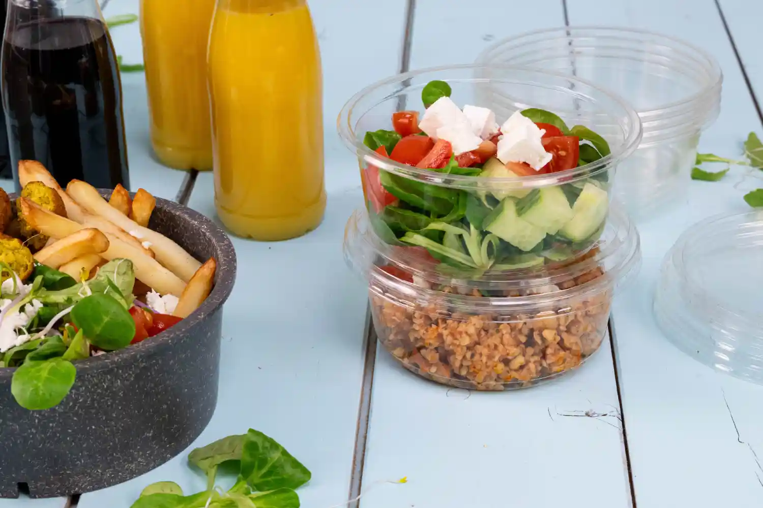 Containers holding salad and grains beside a bowl of fries; two beverage bottles in the background; light blue wooden surface.