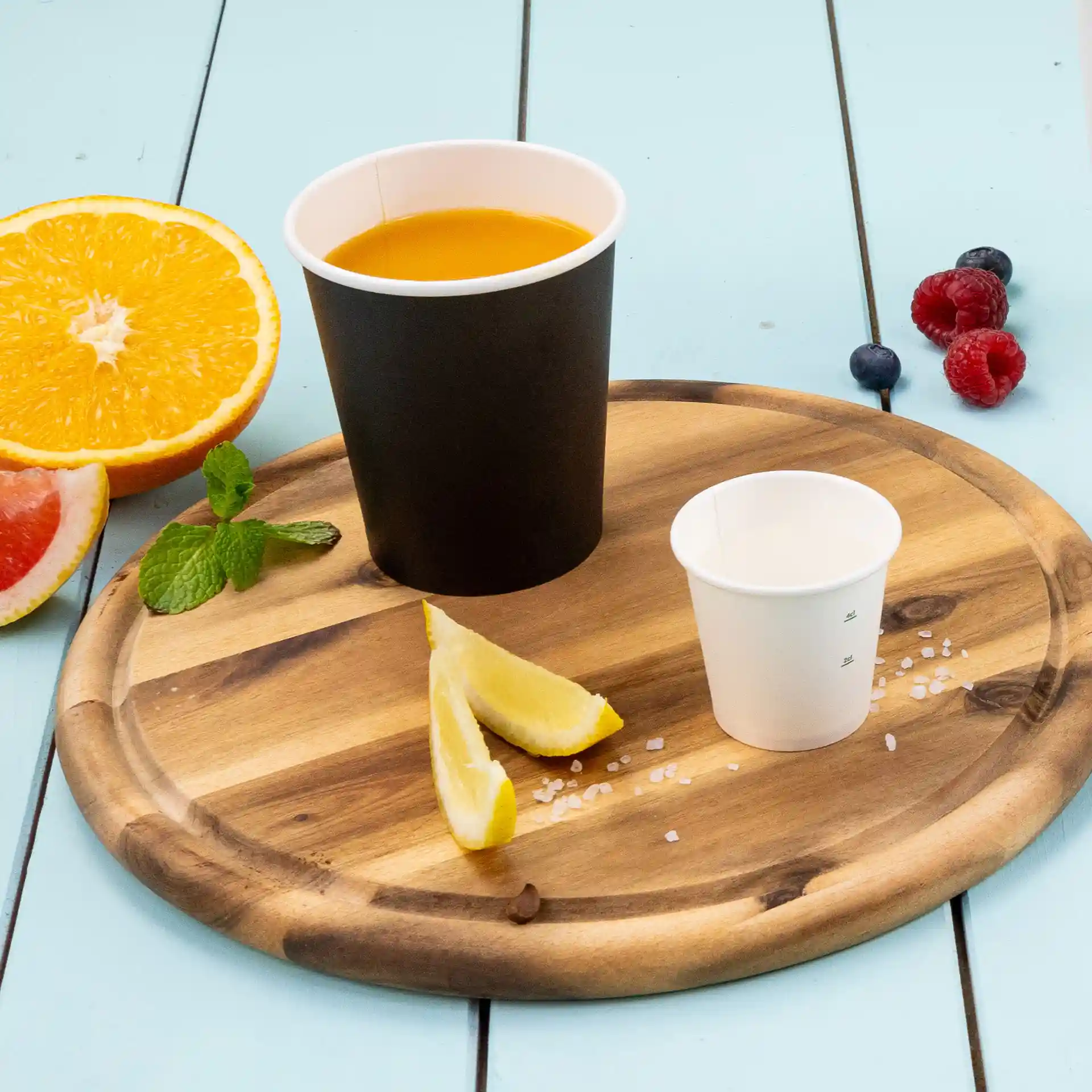 Black paper cup with orange liquid, small white cup, lemon wedges, mint leaves, and fruits on a wooden tray.