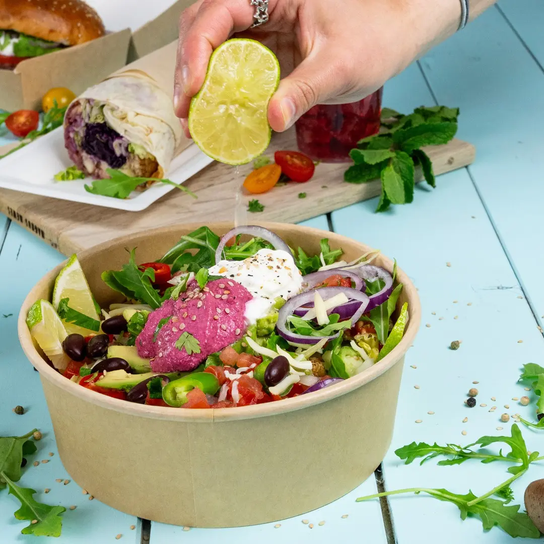 A hand squeezes a lime over a salad in a brown bowl, featuring greens, tomatoes, onions, and pink hummus.