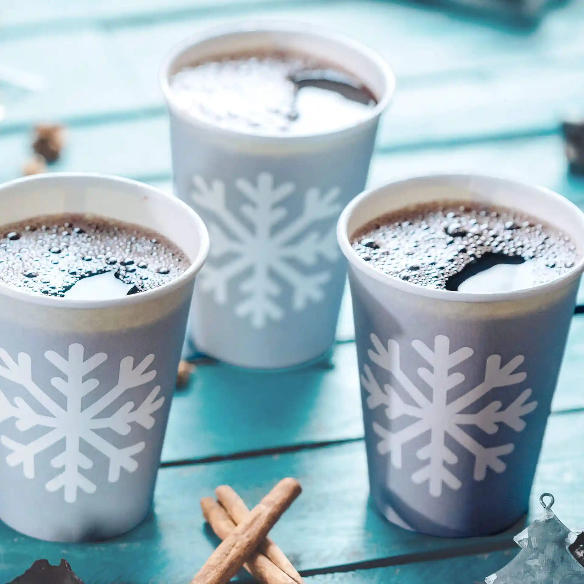 Three grey paper cups with white snowflake designs, filled with dark liquid, on a turquoise wooden surface.