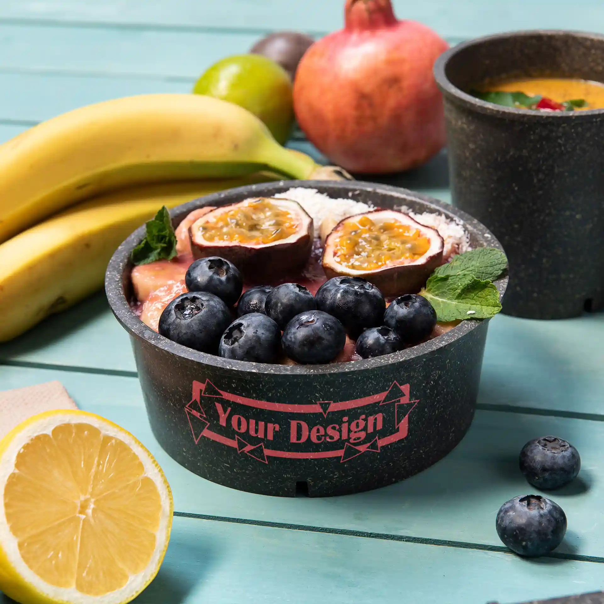 Round grey reusable bowl with fruit toppings, including blueberries and passion fruit, on a teal surface.
