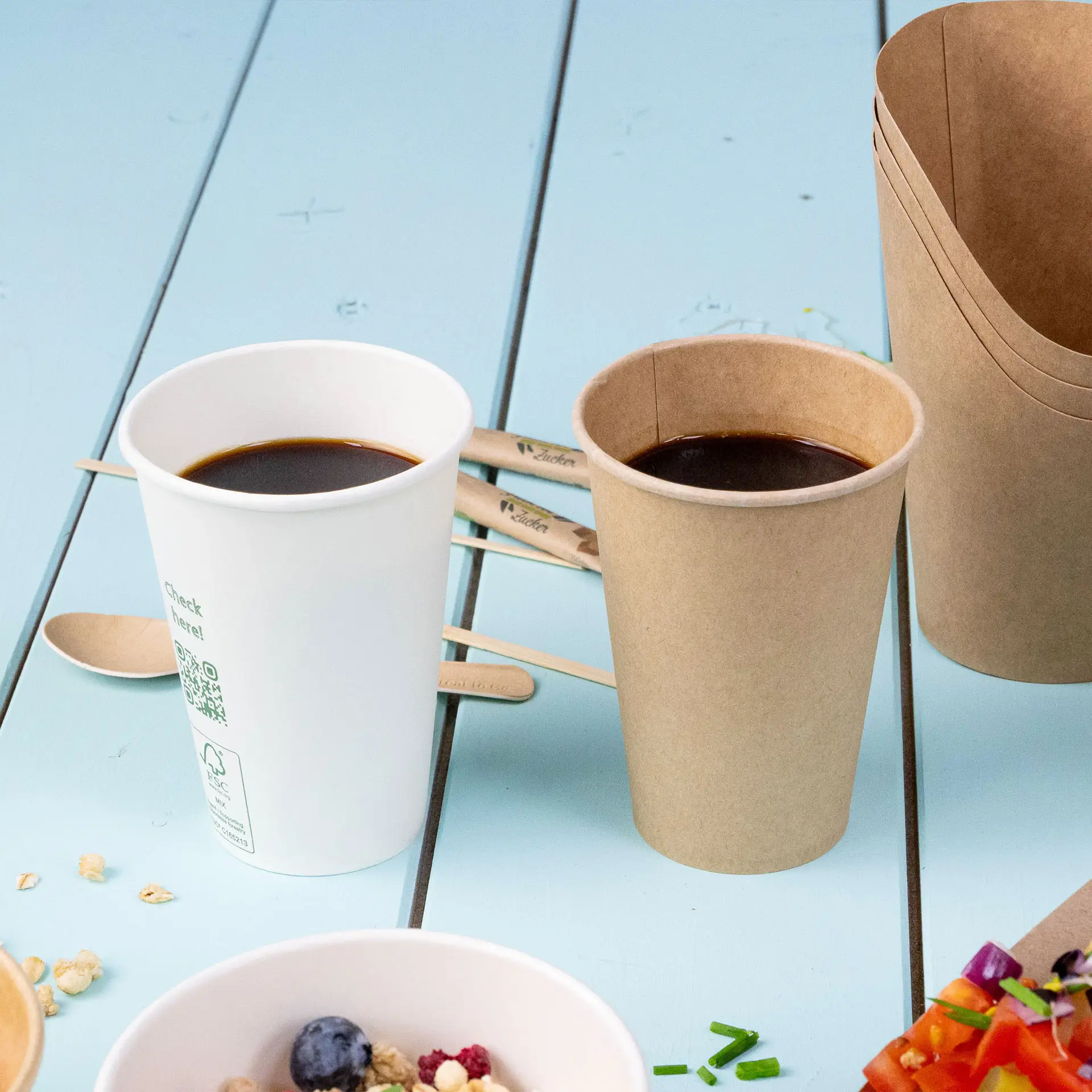 One white paper coffee cup and one brown paper cup filled with dark liquid; wooden stirrers in the background.