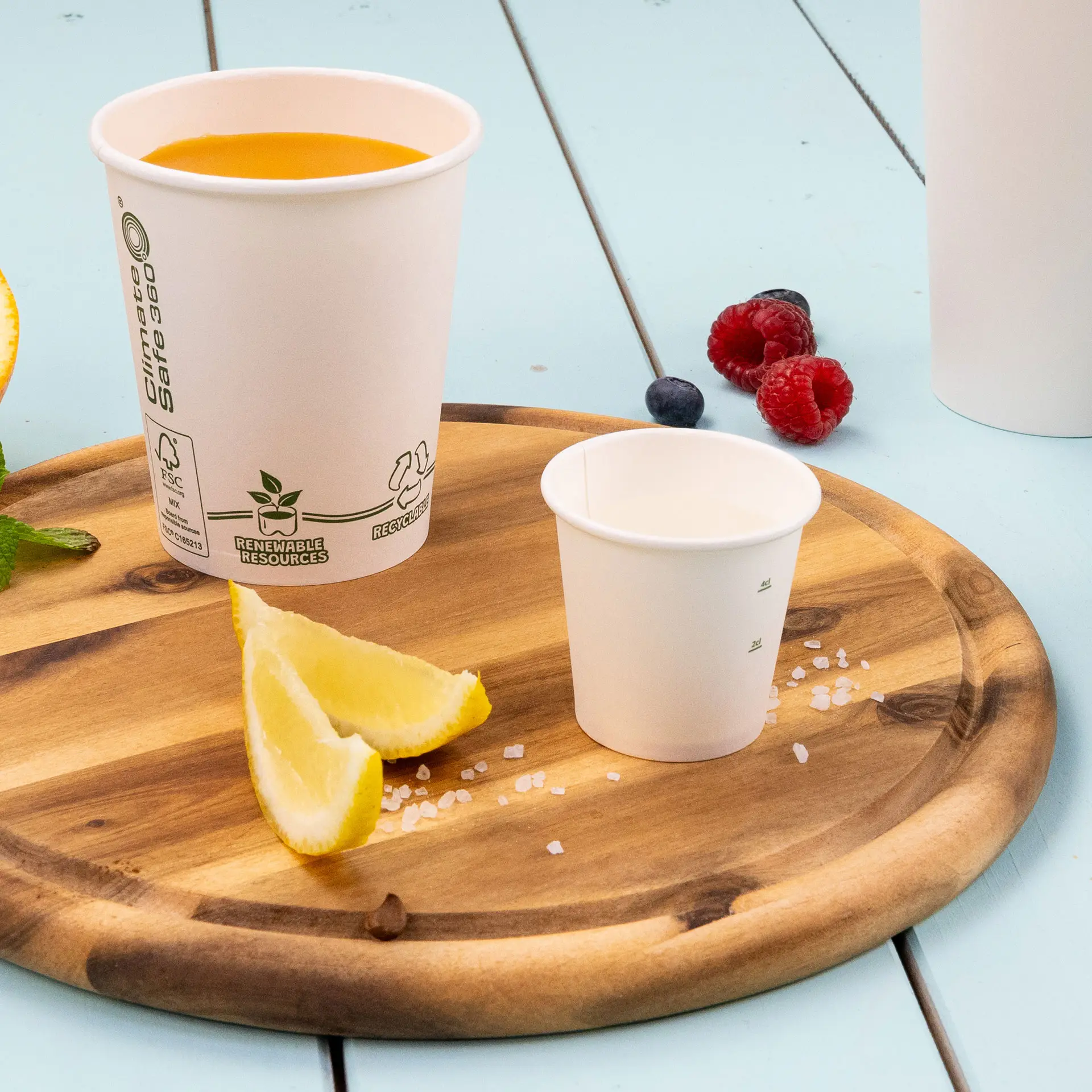 Single-wall white paper cup, 50 ml, PLA-coated for hot and cold drinks; surrounded by a small cup, lemon wedges, and berries on a wooden tray.