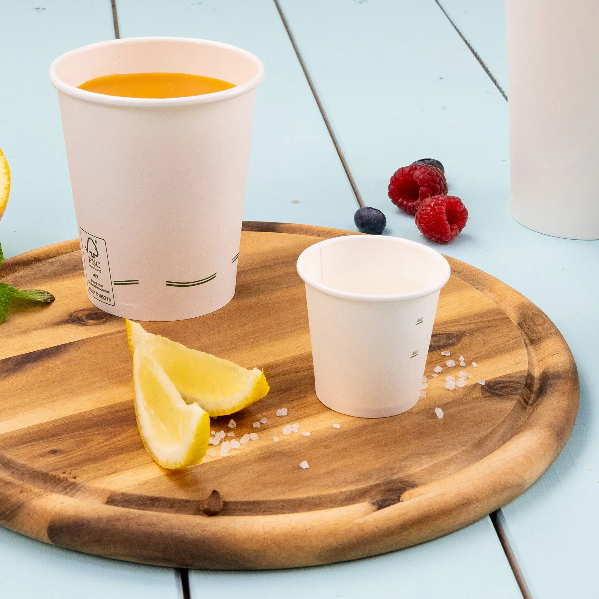 Image of a wooden platter with a larger white paper cup filled with orange liquid, a smaller white paper cup, lemon wedges, and scattered salt.