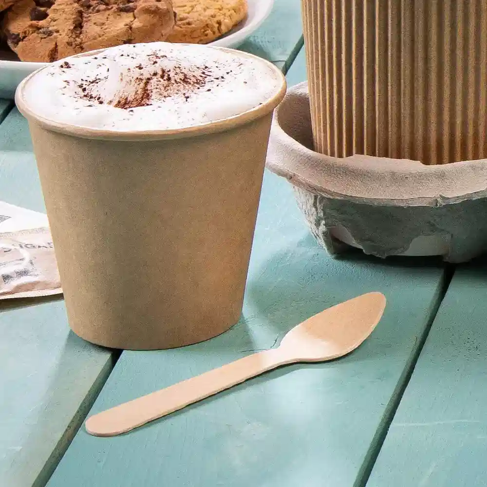 Brown disposable cup with foam topping and a wooden spoon on a teal table, accompanied by cookies in the background.