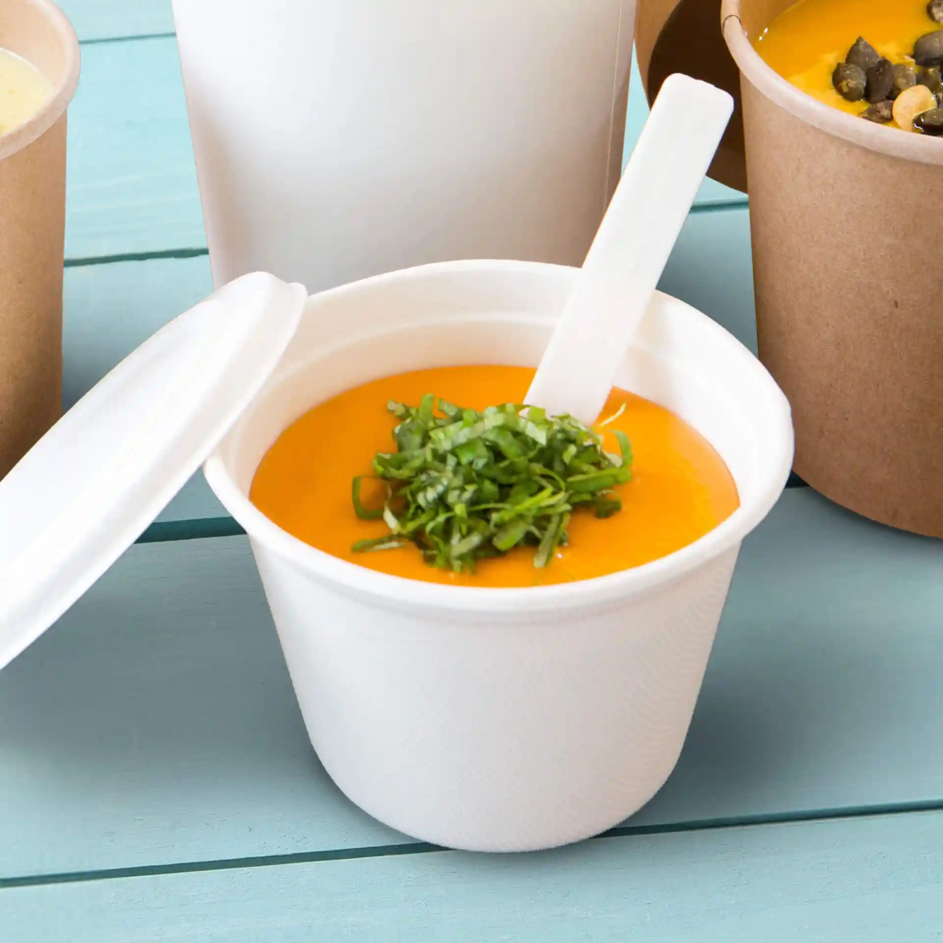 White sugarcane soup cup filled with orange soup and topped with chopped green herbs, with a white spoon inside.