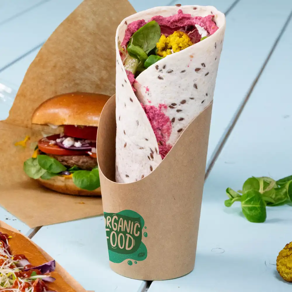 A wrapped flatbread filled with colorful vegetables, displayed in a kraft paper holder with a label reading "Organic Food", alongside a burger and salad.