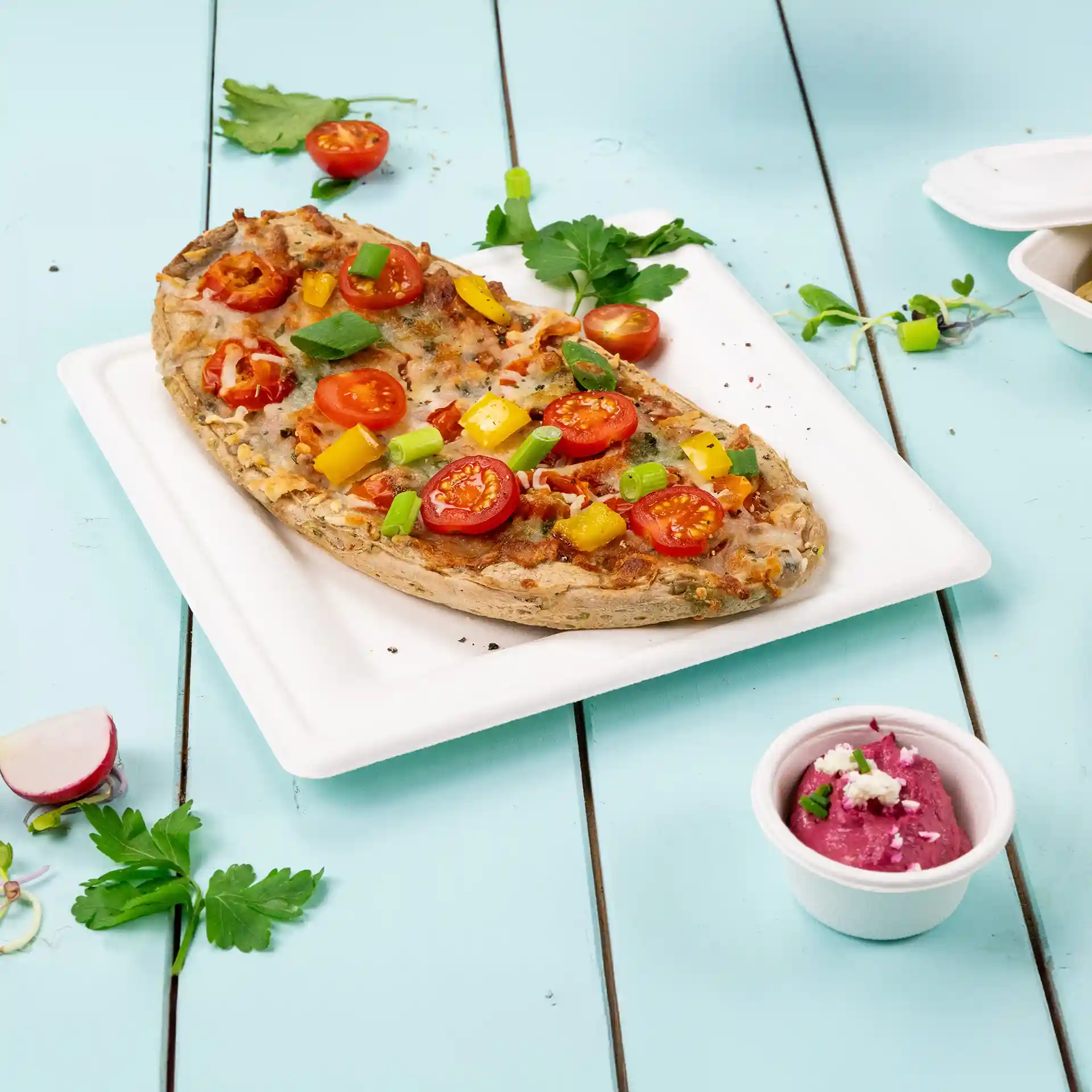 Square white plate made of sugarcane with a pizza topped with tomatoes, peppers, and green onions, alongside a small cup of dip.