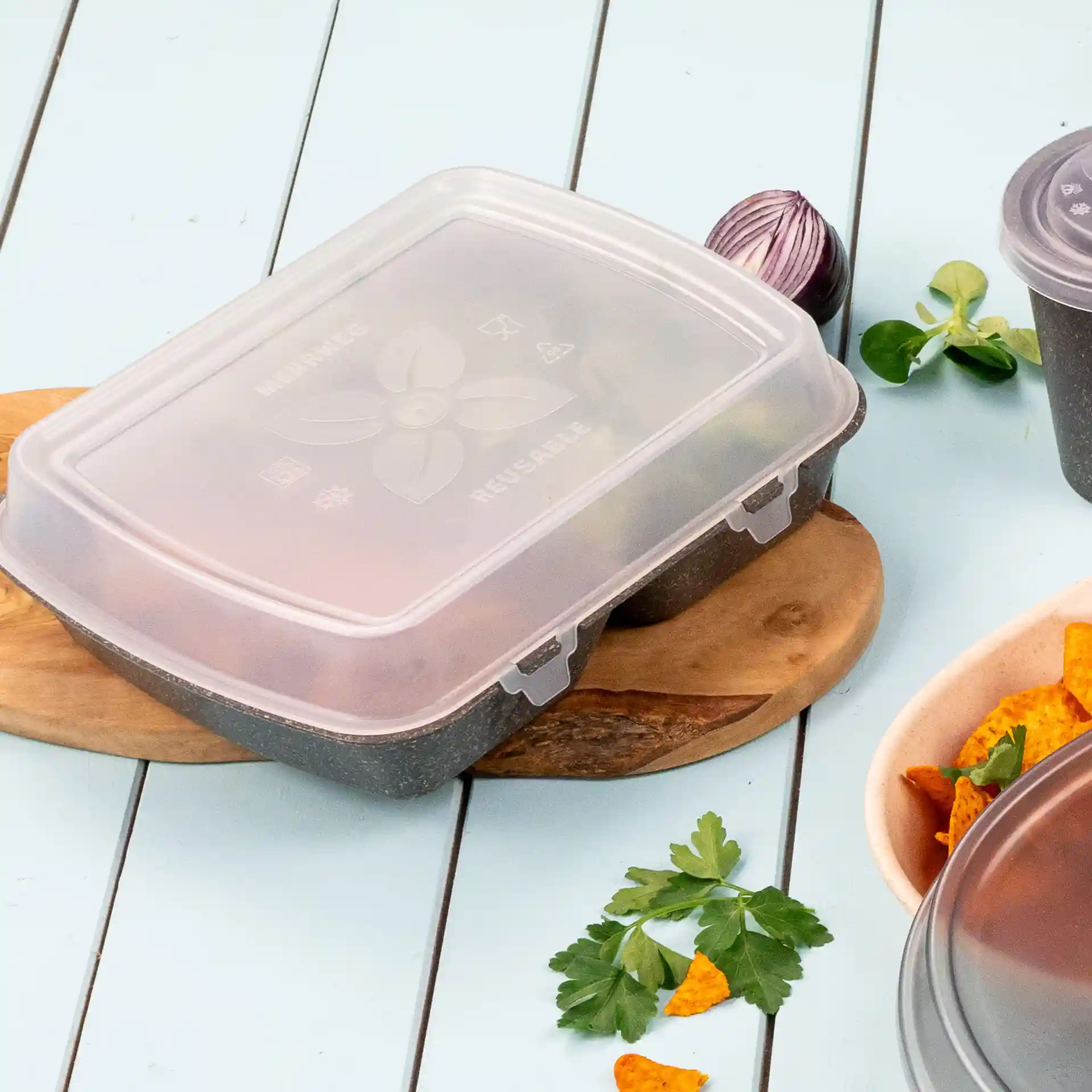 Transparent reusable food container with a lid, placed on a wooden board, surrounded by herbs and snacks.