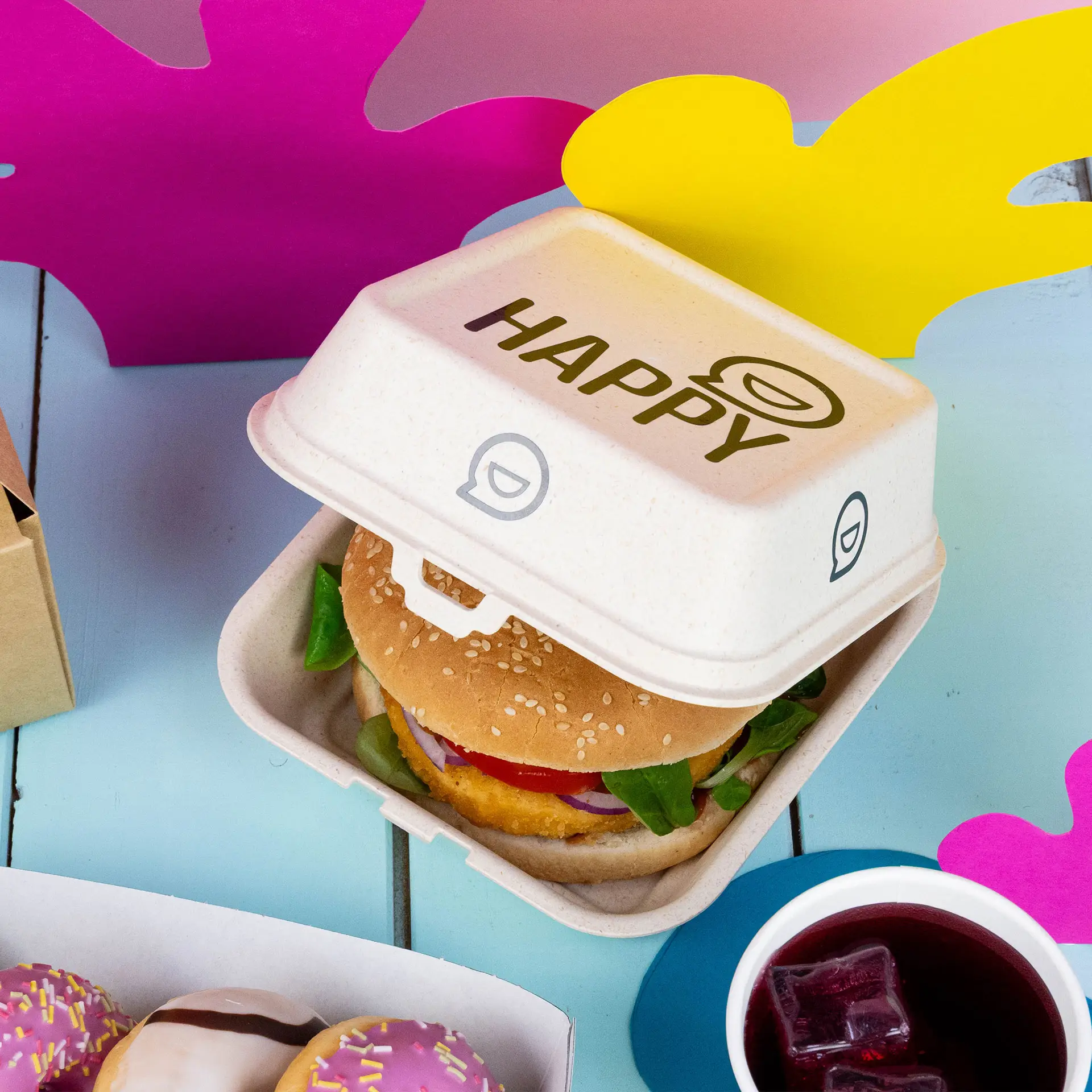 A cream white reusable burger box with an orange logo label, suitable for hot and cold foods, on a table next to a burger.