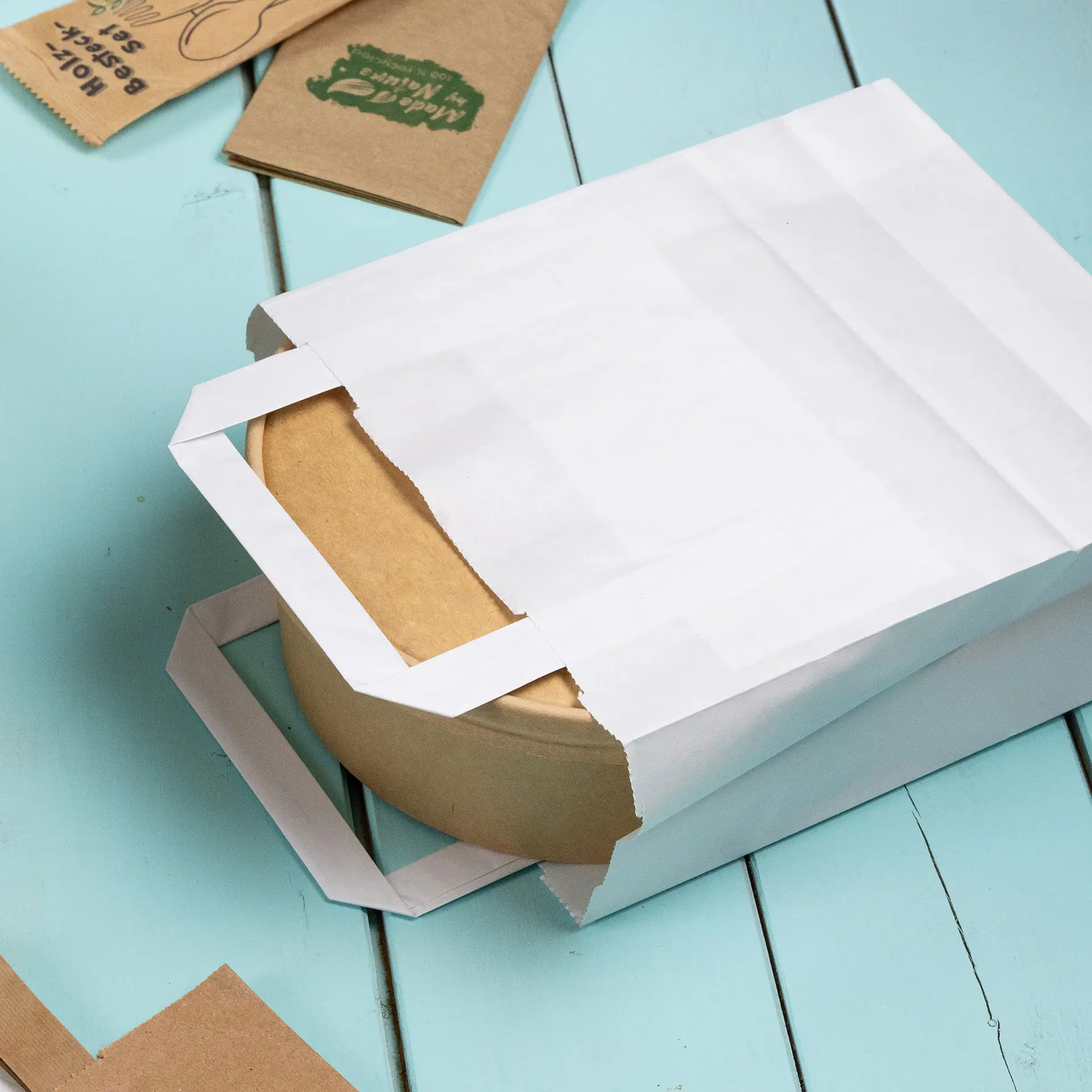 White paper carrier bag partially opened, revealing a round kraft paper container inside on a light blue surface.