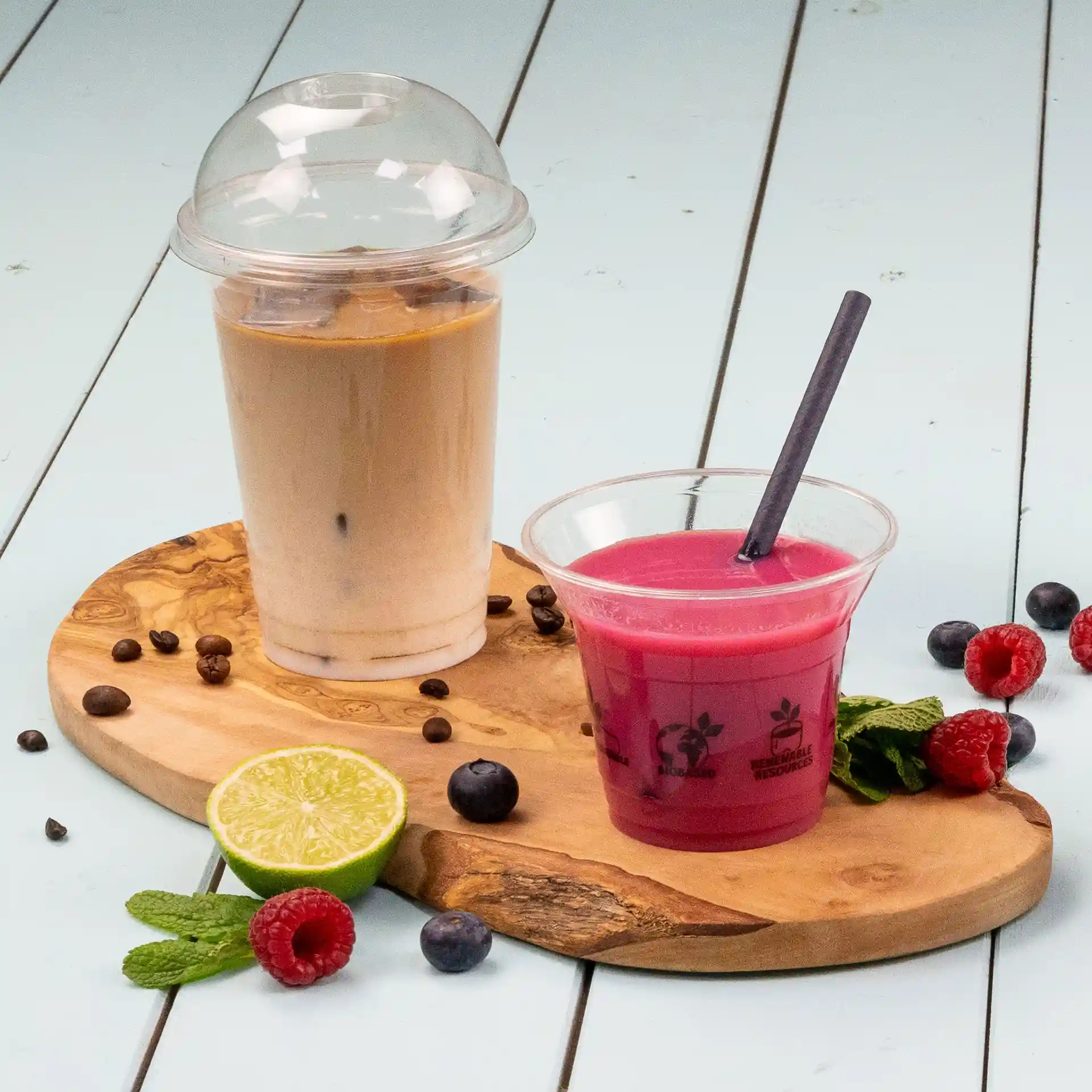 Image of a transparent cup with a dome lid filled with a light brown beverage, next to a pink drink in a clear cup with a straw, on a wooden board with fruits.