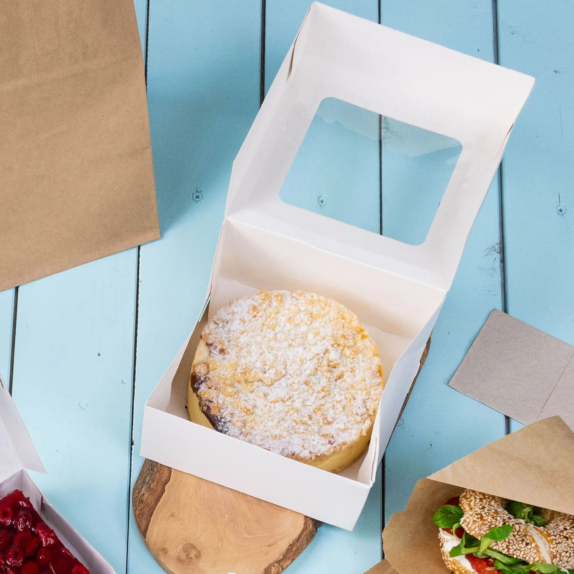 Cake box M in white with an open lid showing a round cake inside, placed on a wooden surface with other food items nearby.