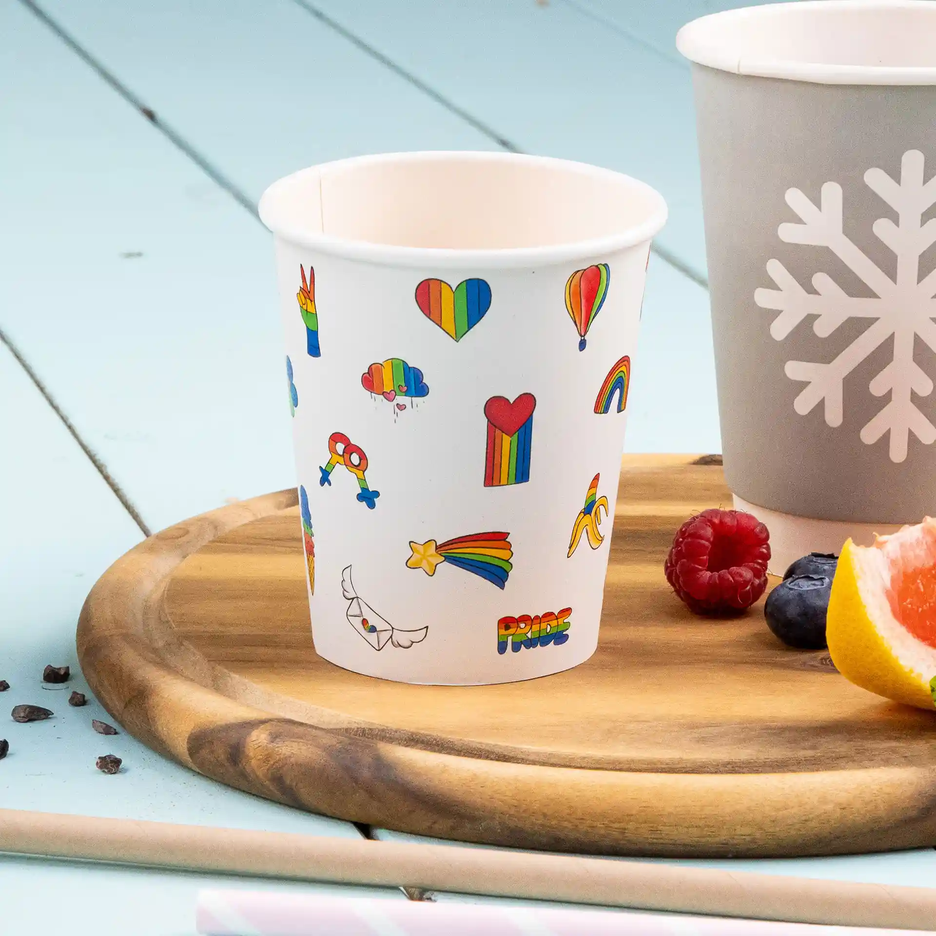White paper cup with colorful printed designs, including hearts, rainbows, and clouds, on a wooden tray.