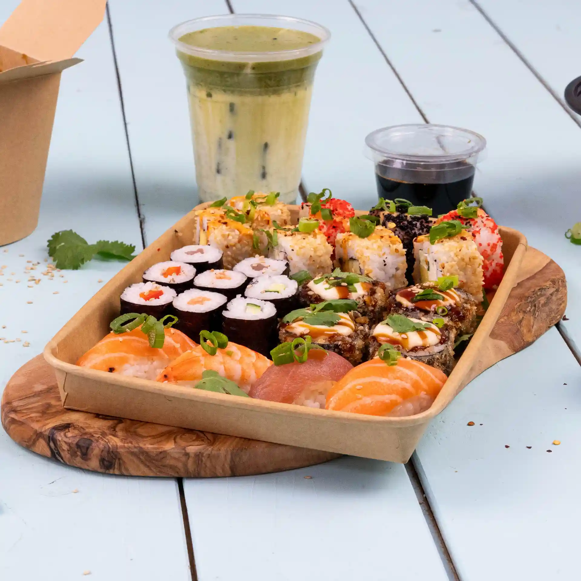 Tray of assorted sushi rolls, including shrimp and salmon, with a drink and dipping sauce on a wooden board.