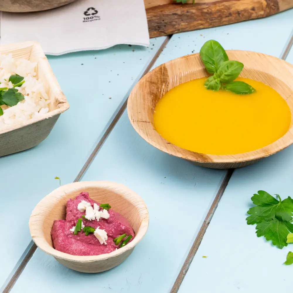 Bamboo bowl filled with yellow soup garnished with a basil leaf; smaller bowl with pink dip topped with herbs; rice with herbs in a container nearby.